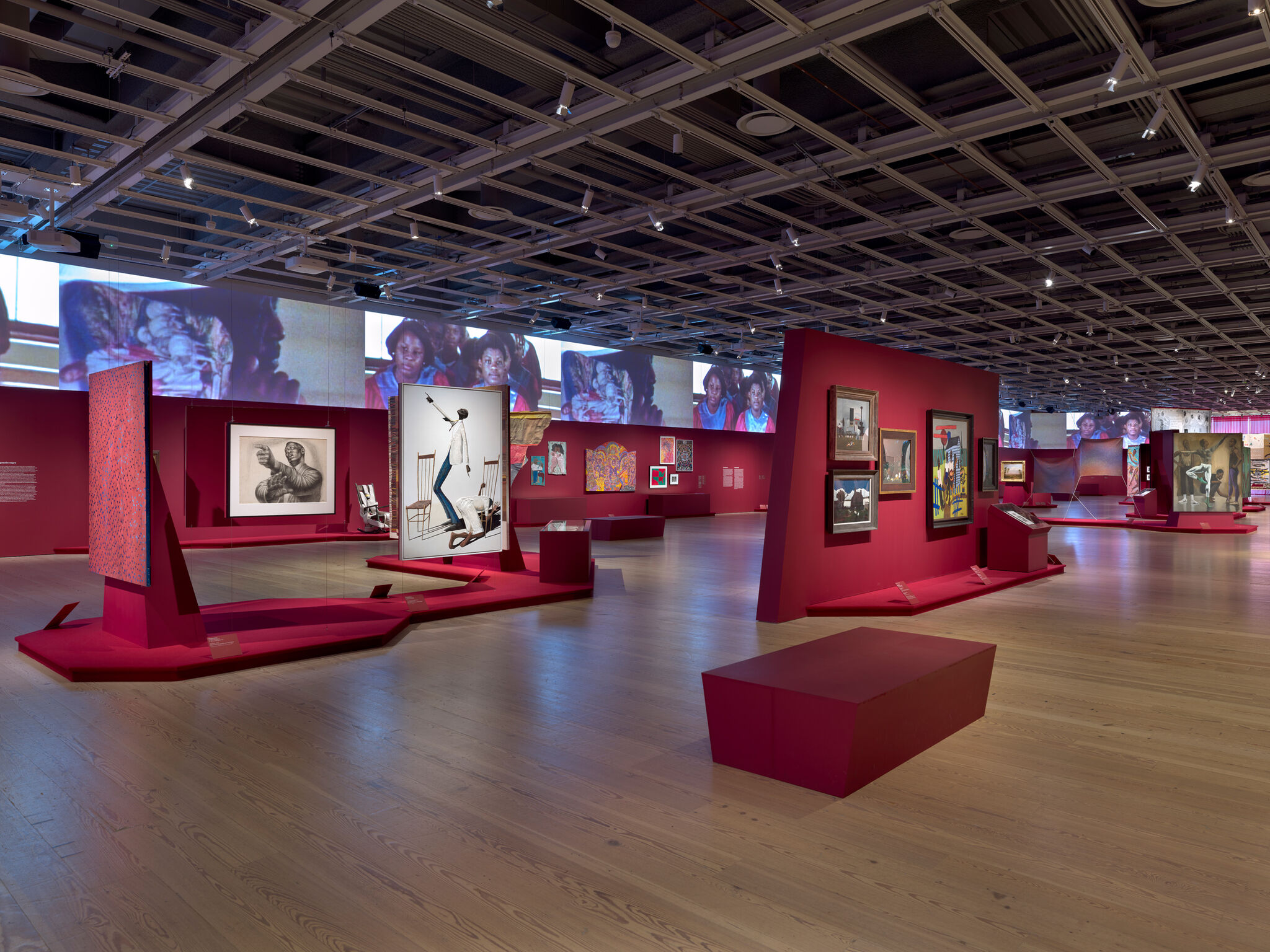 Art gallery with red walls displaying various paintings and photographs. Large screens above show images of people. Wooden floor and modern design.