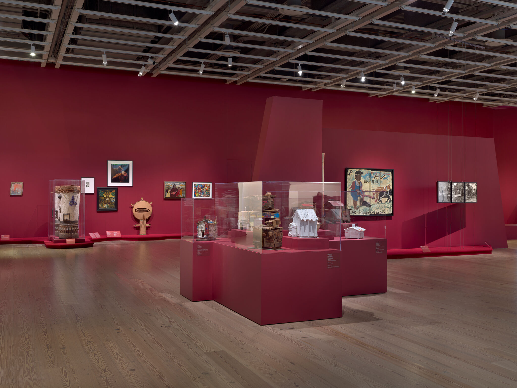 Art gallery with red walls displaying various paintings and sculptures in glass cases. Wooden floor and overhead lighting.