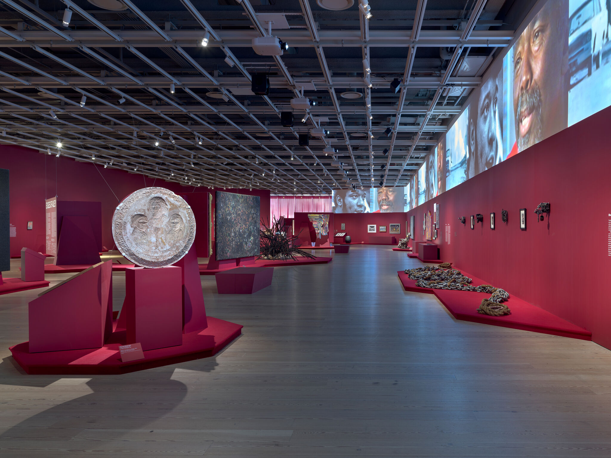 Art gallery with red walls, featuring a large circular sculpture, various artworks, and projected images of faces on the upper wall.