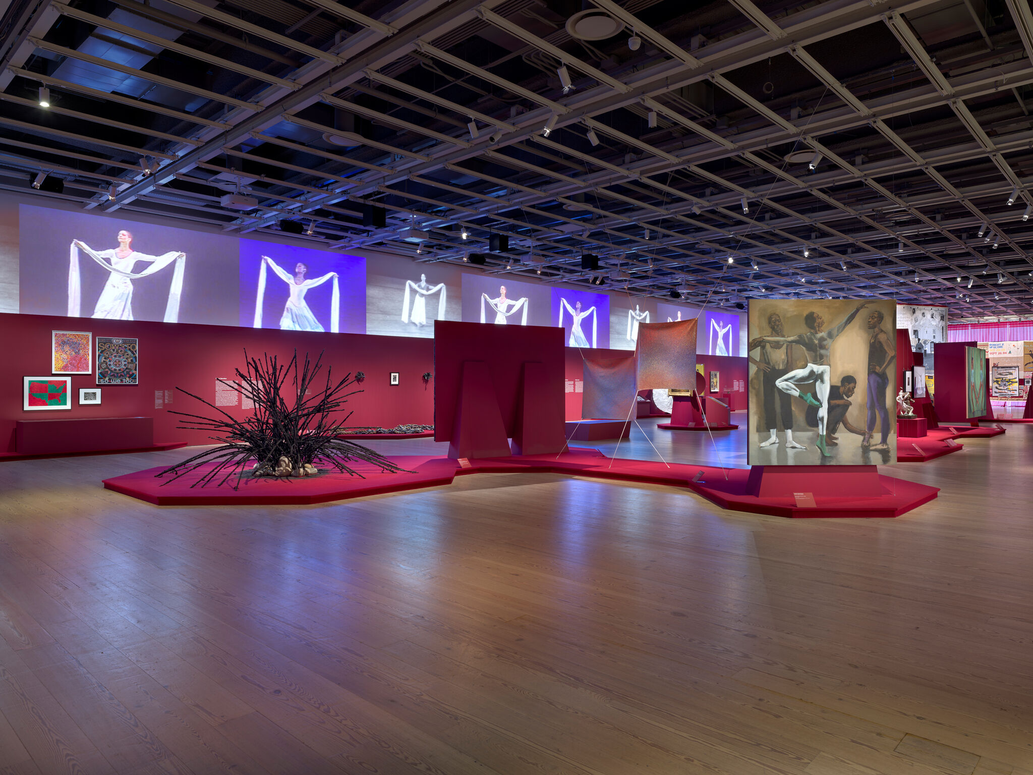 Art gallery with vibrant red walls, featuring sculptures, paintings, and video projections. A central black sculpture stands out on a red platform.