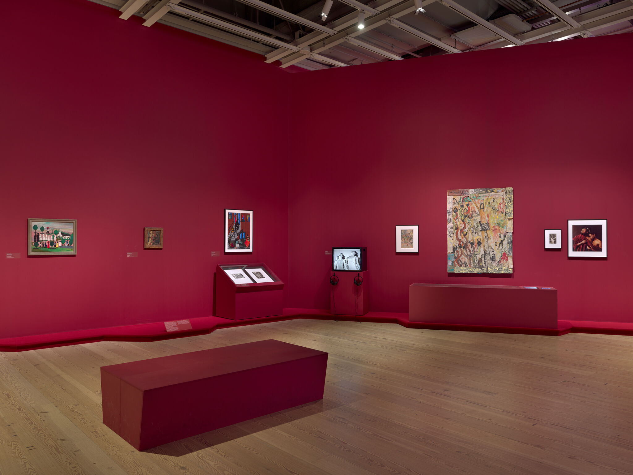 Art gallery with red walls displaying various paintings and a video screen. A red bench sits on the wooden floor.