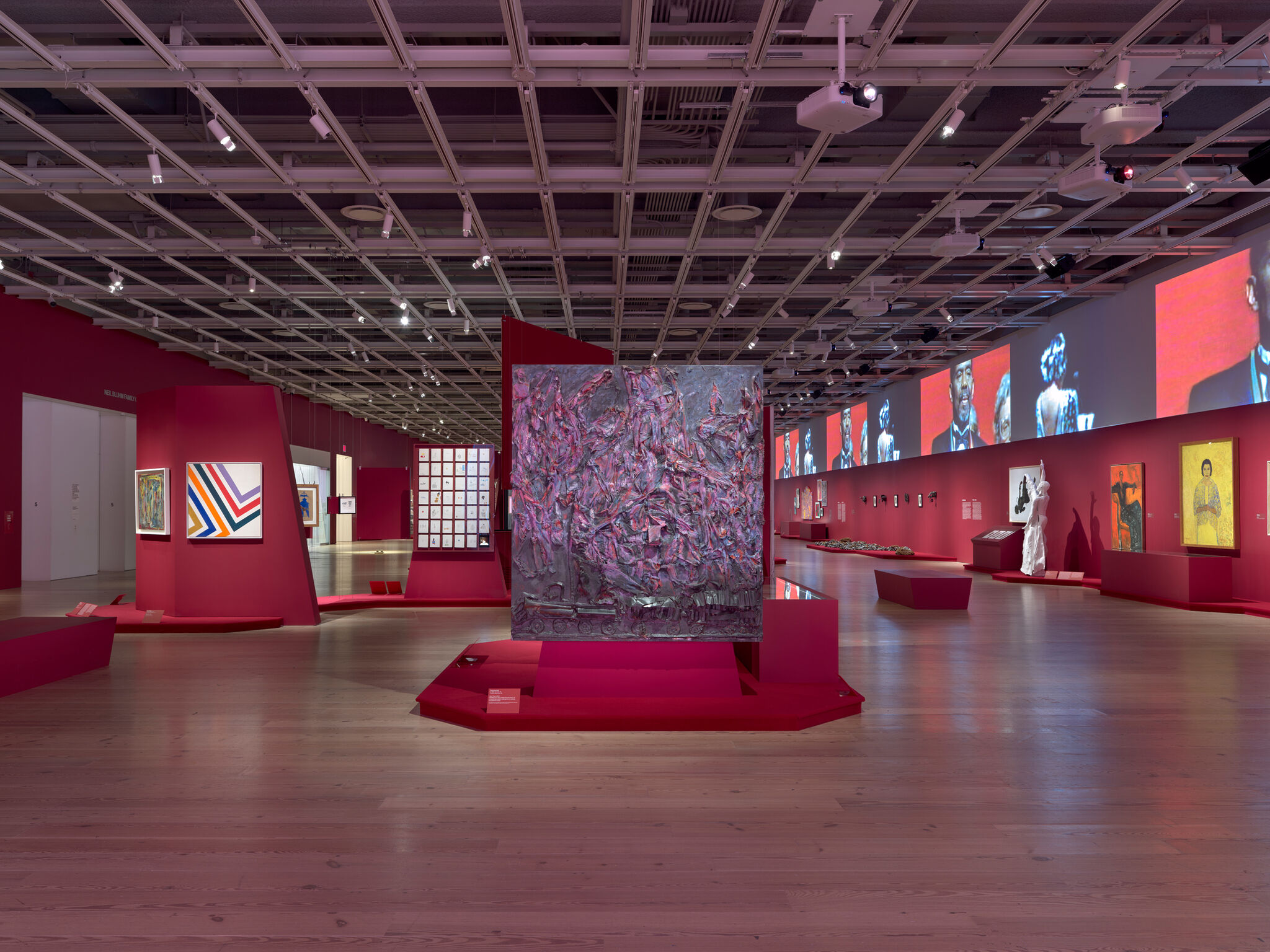 Art gallery with vibrant red walls, featuring abstract paintings and sculptures. A large textured artwork is centered, with digital displays above.