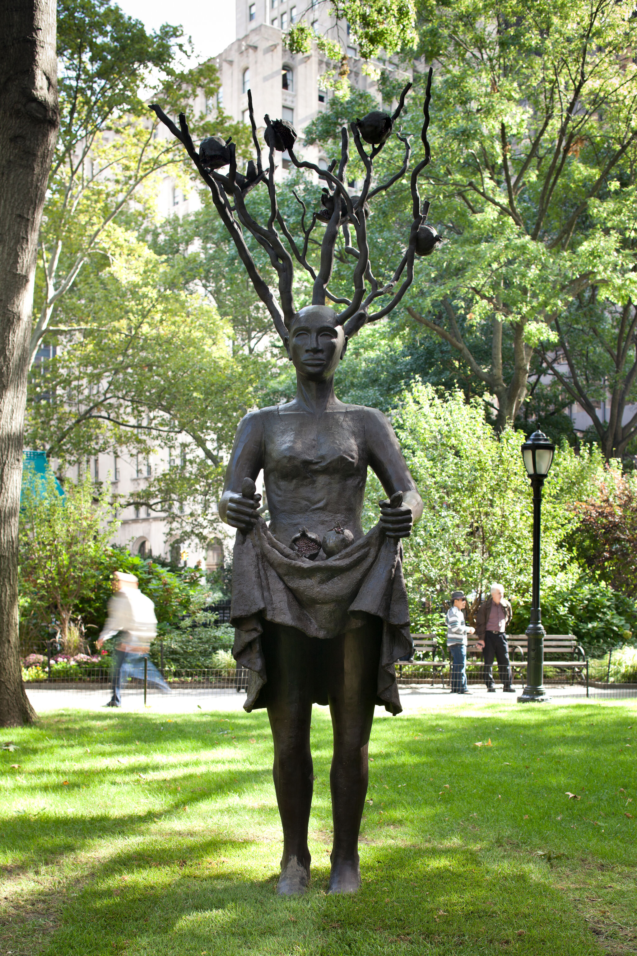 Sculpture of a woman with tree branches for hair, set in a park with trees and buildings in the background.