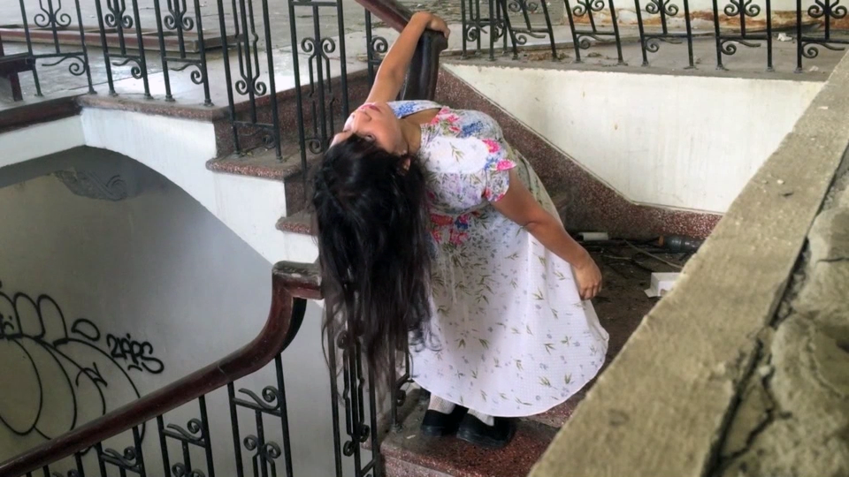 A woman in a floral dress leans back over a stair railing in an old building with graffiti on the walls.