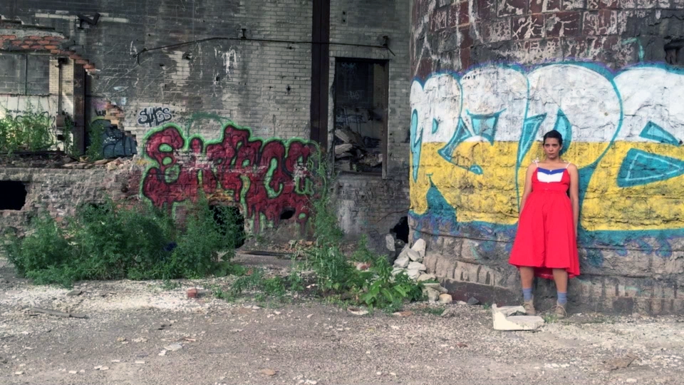 A person in a red dress stands against a graffiti-covered wall in an abandoned, overgrown industrial area.