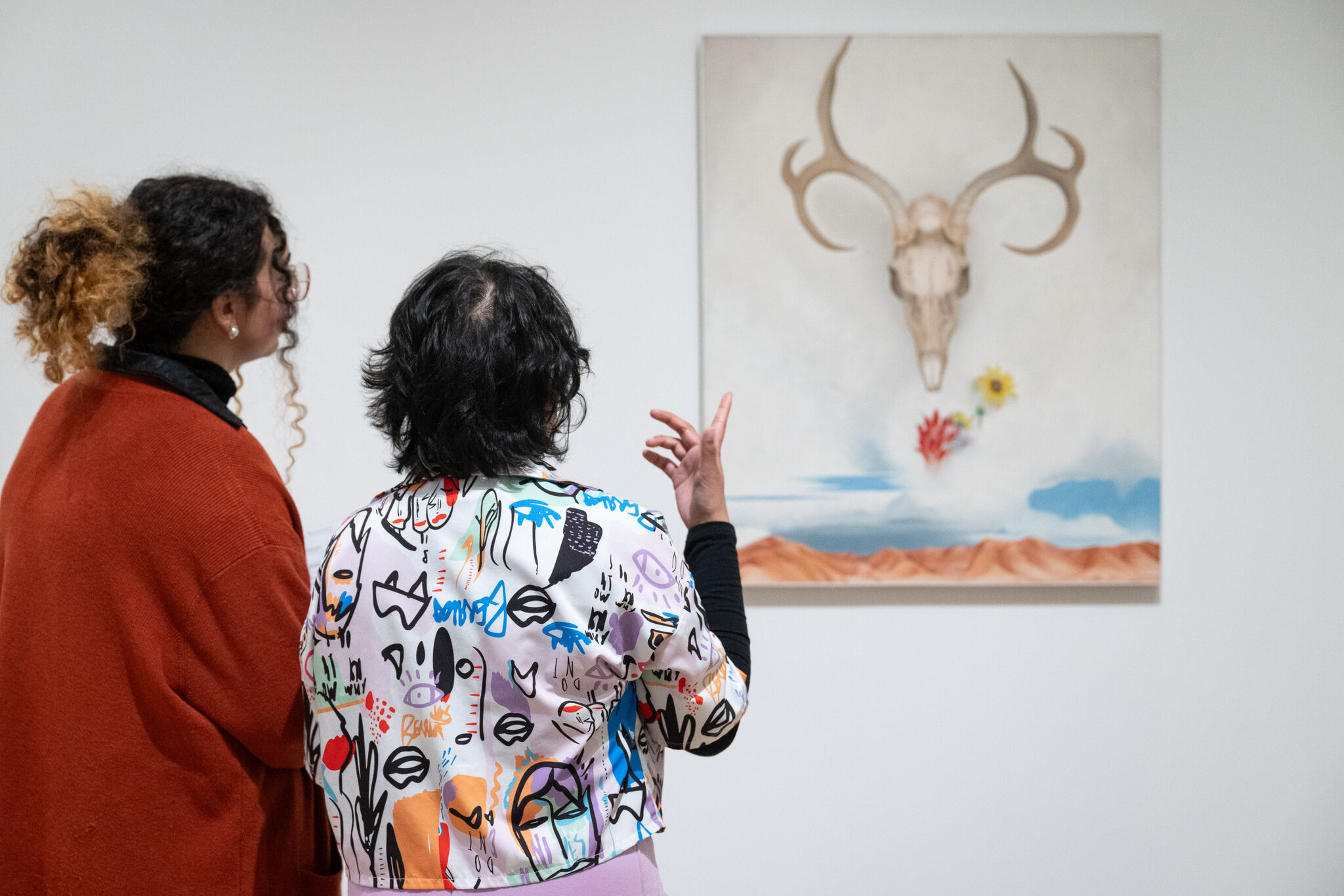 Two people observe a painting of a skull with antlers, flowers, and a desert landscape. One gestures while discussing the artwork.