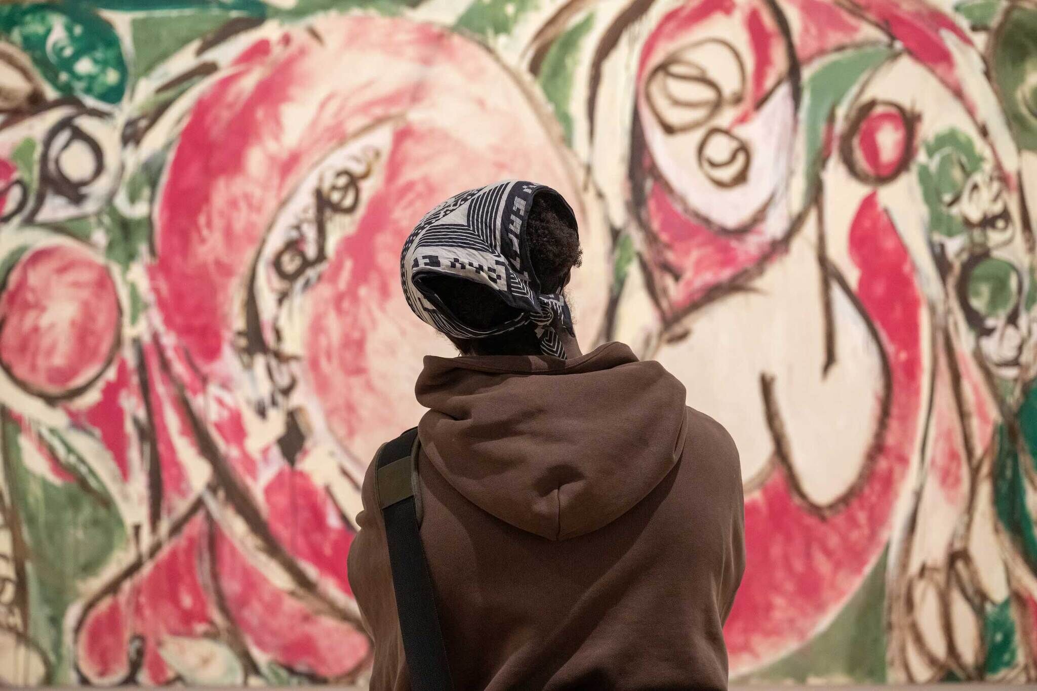 Person in a brown hoodie and patterned headscarf views an abstract painting with red, green, and white swirls in a gallery.