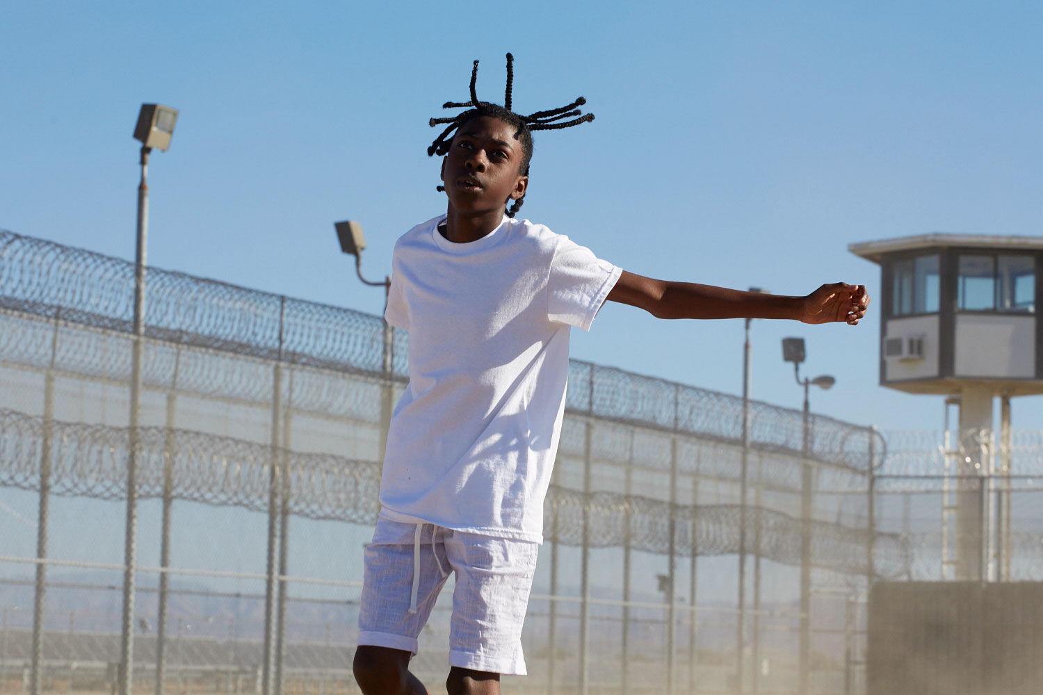 A young black boy wearing all white is photographed hopping mid-air with his arms stretched near the barbed wire fences of a correctional facility