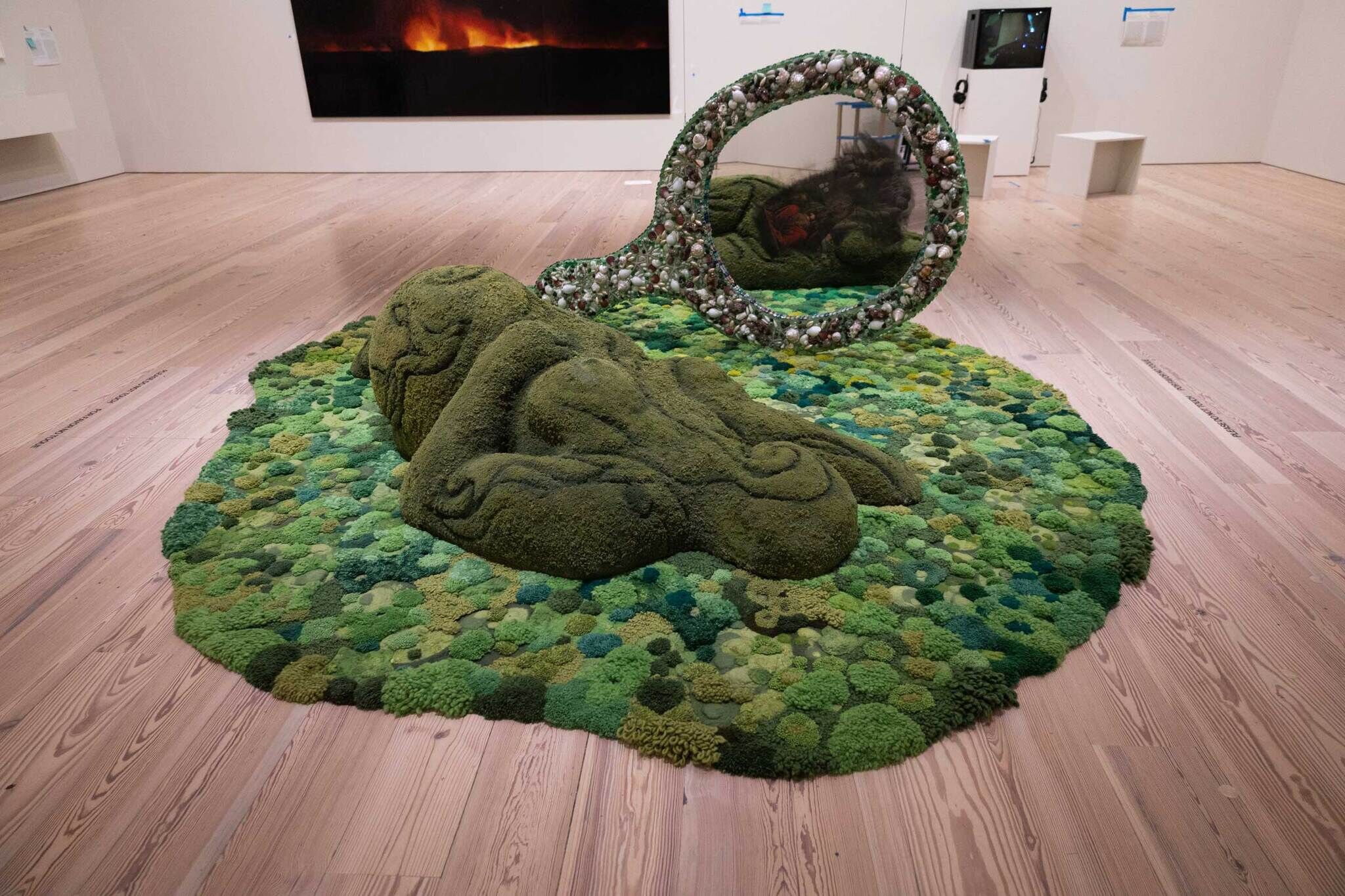 Green moss sculpture resembling a reclining figure with a mirror, displayed on a wooden floor in an art gallery.