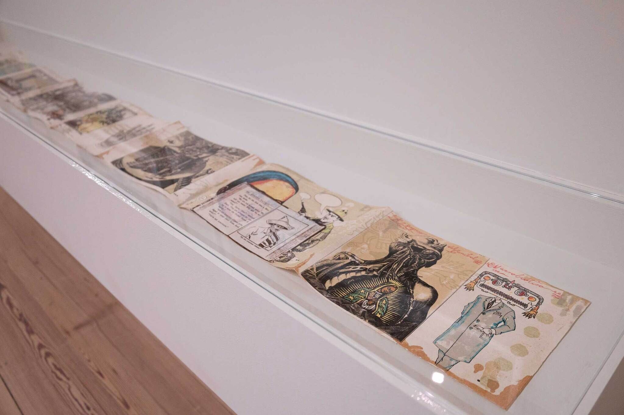 A series of illustrated pages displayed in a glass case, featuring colorful, abstract designs and text on a wooden floor.