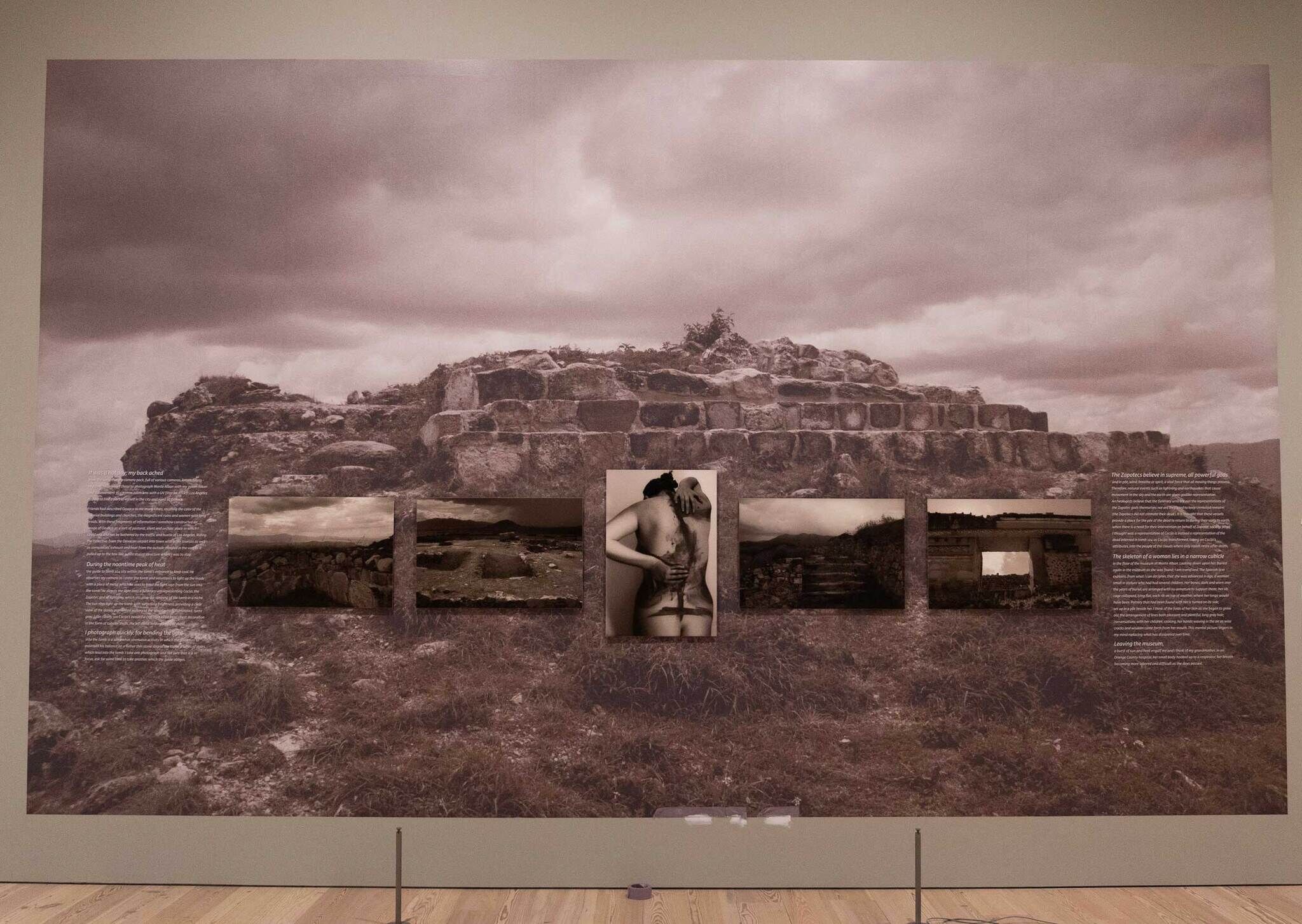 A large sepia-toned photograph of ancient stone ruins with smaller images and text panels displayed below it on a gallery wall.
