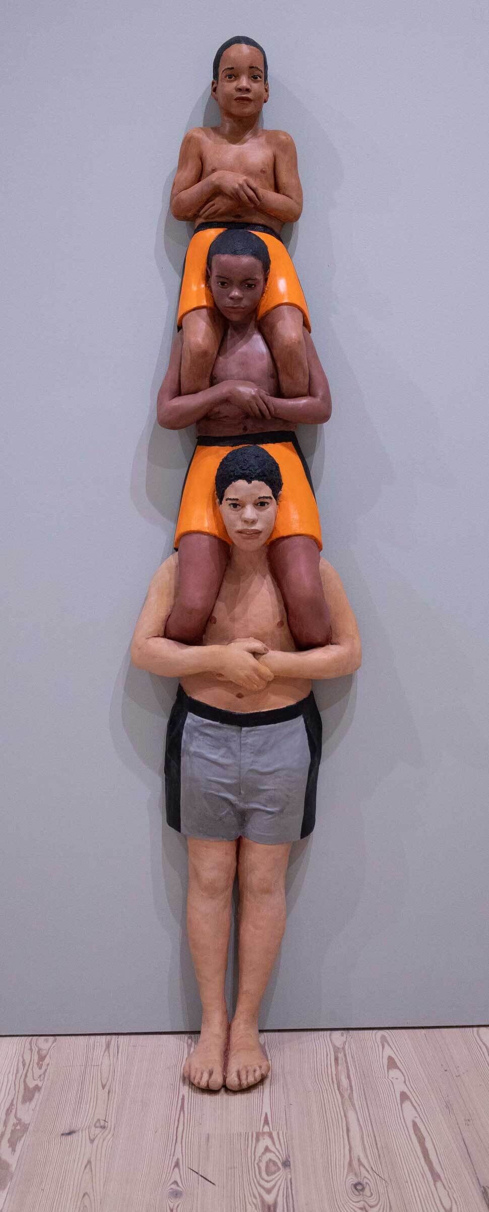 Sculpture of three boys stacked on each other's shoulders, wearing orange and gray shorts, against a plain background.