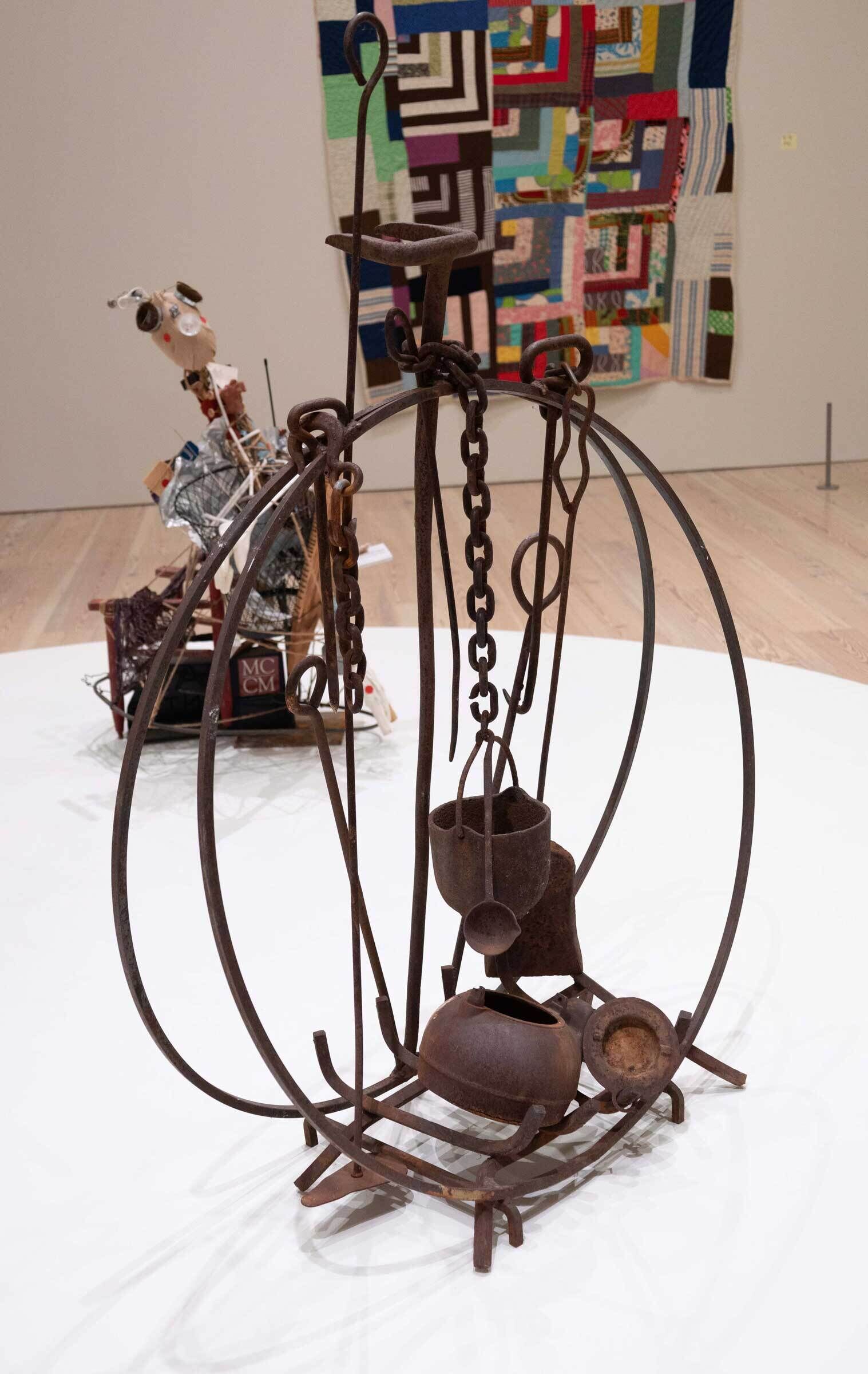 Rustic metal sculpture with chains and pots in the foreground, colorful patchwork quilt on the wall, abstract art piece in the background.