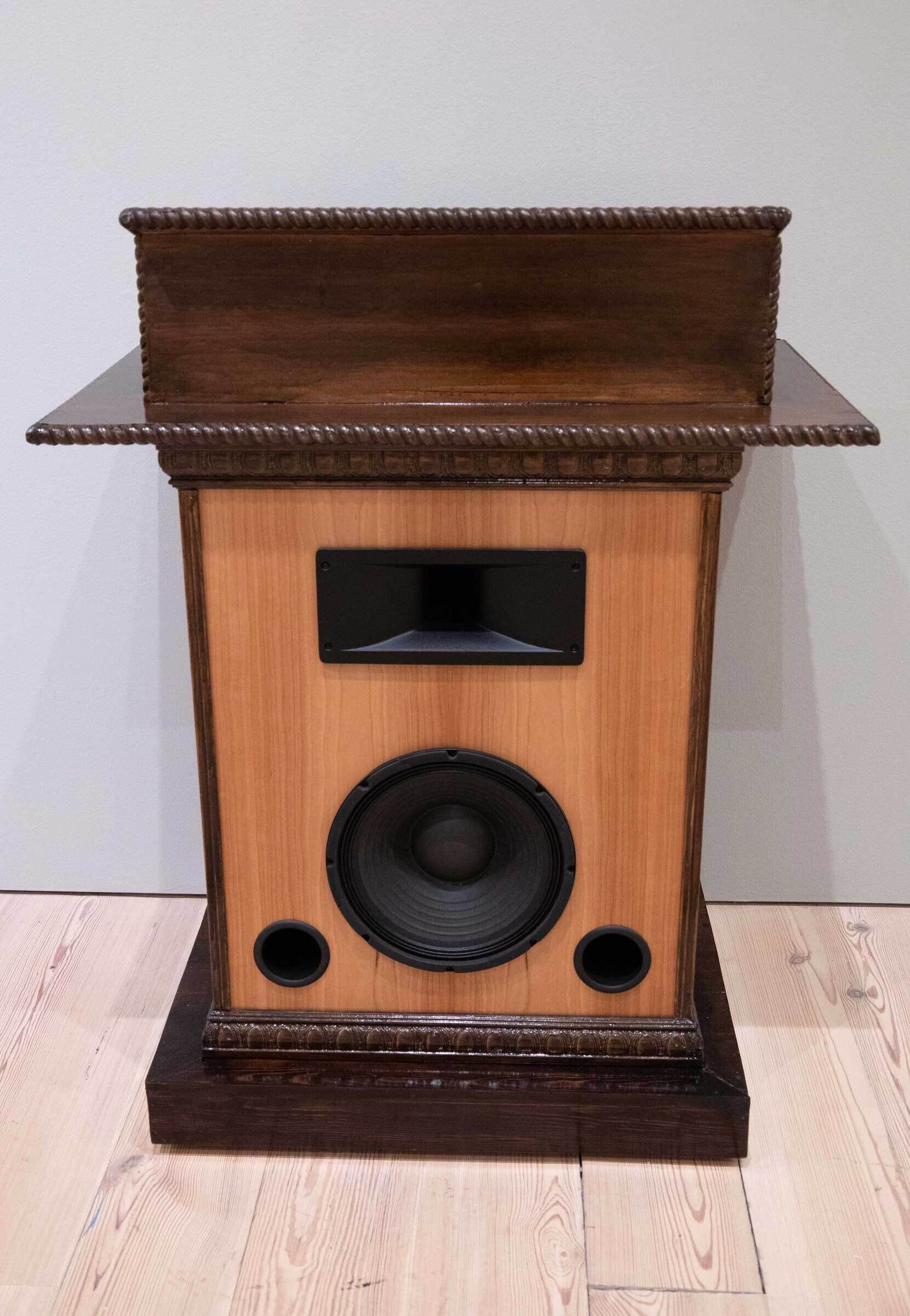 A vintage wooden speaker with ornate detailing, featuring a large woofer and two smaller ports on a wooden floor.