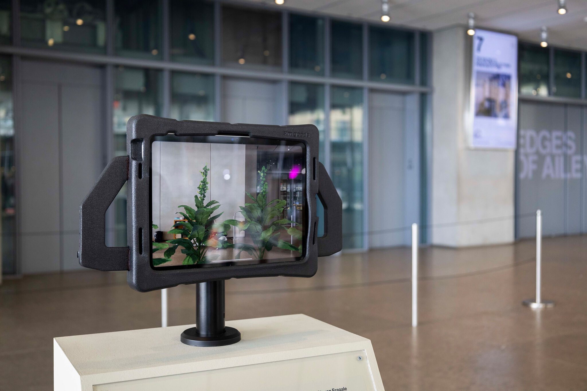 A digital display shows plants in a modern indoor setting, mounted on a stand in front of a glass-walled room.