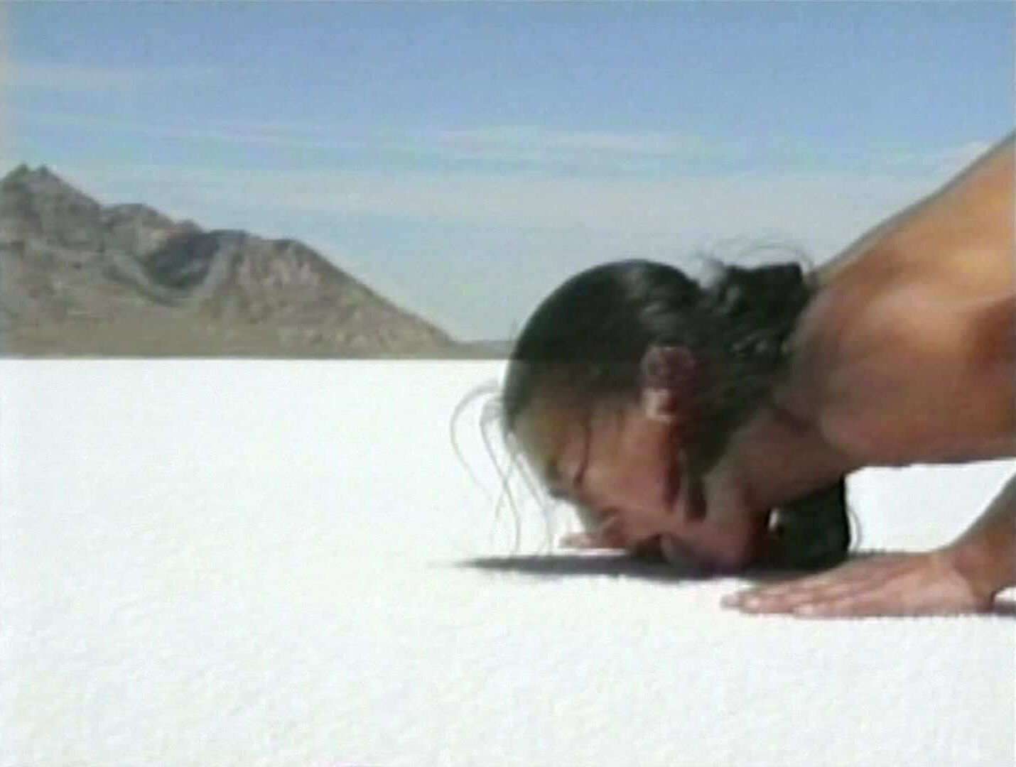 Person with long hair lying face down on a vast white salt flat, with distant mountains under a clear blue sky.