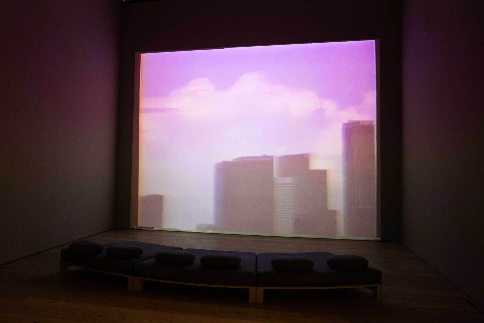 A dimly lit room with a large screen displaying a blurred cityscape under a purple sky, and a curved bench with cushions in the foreground.