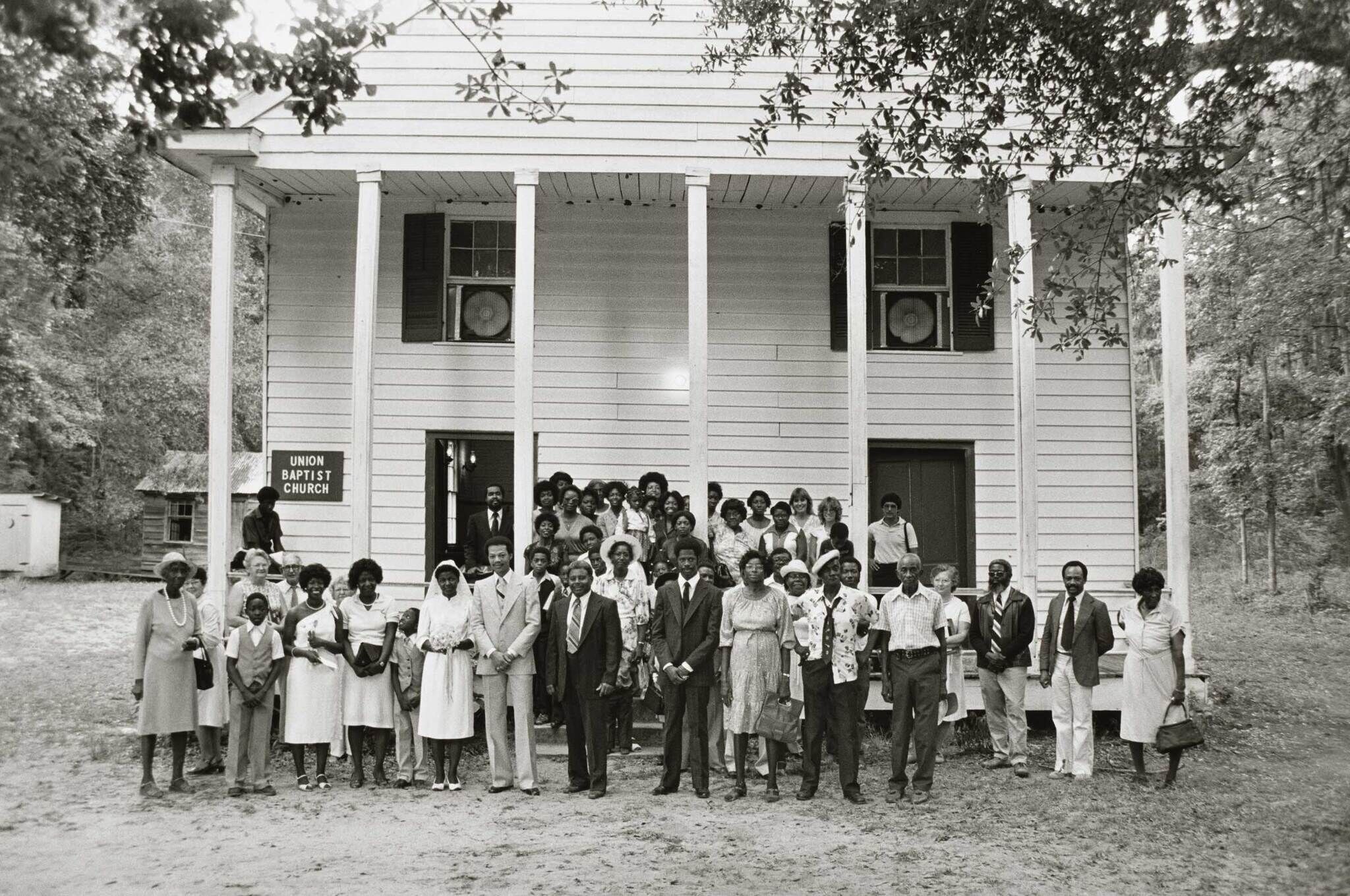 A group of people stands in front of Union Baptist Church, a white wooden building with tall columns, surrounded by trees.