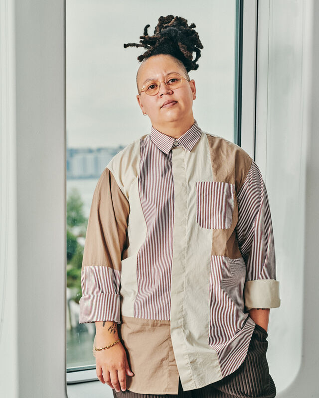 Person with glasses and dreadlocks stands by a window, wearing a striped and patchwork shirt, with a cityscape in the background.