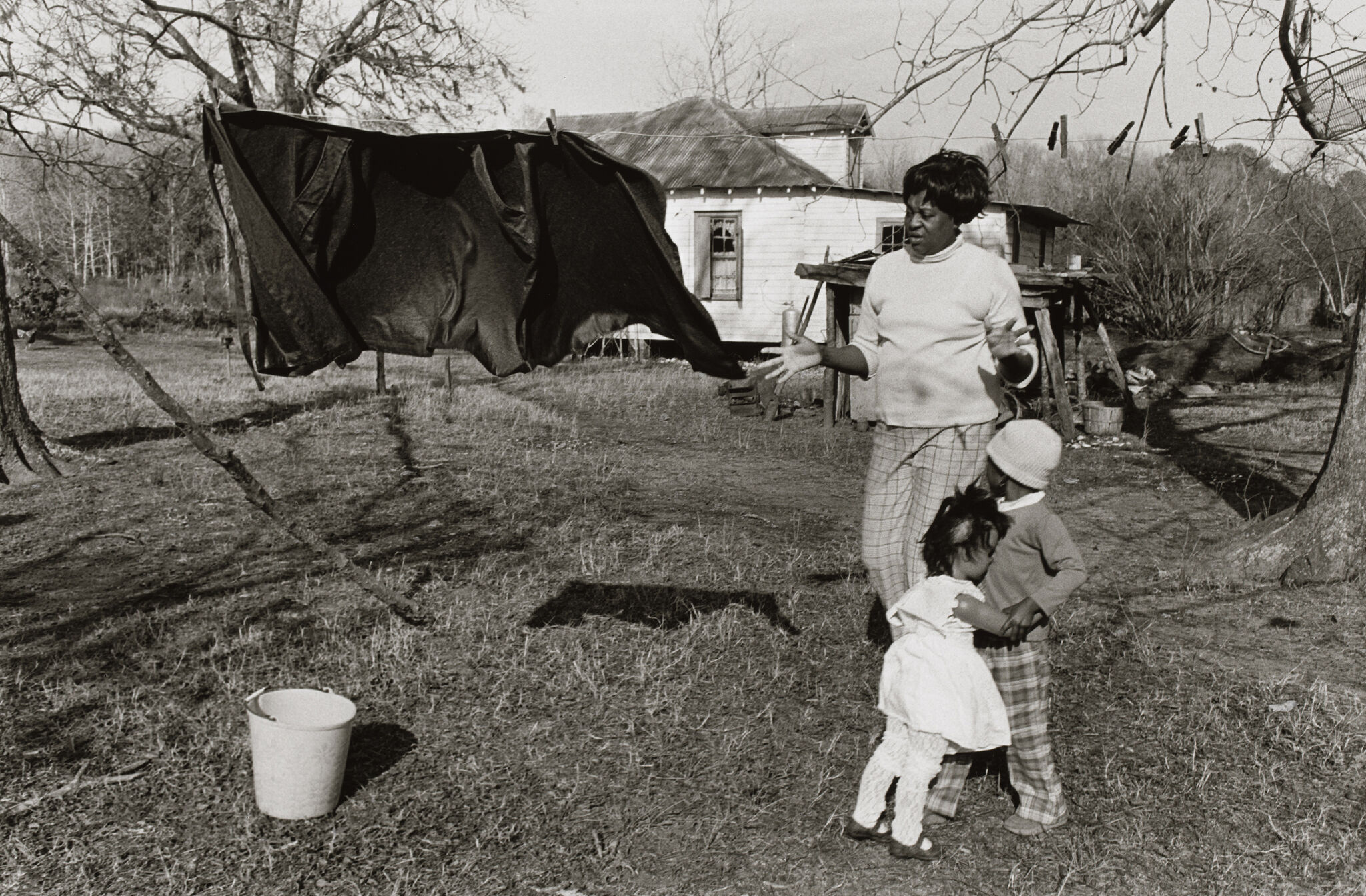 A woman hangs laundry on a line outside a rural house while two children hug nearby. A bucket sits on the ground in the foreground.