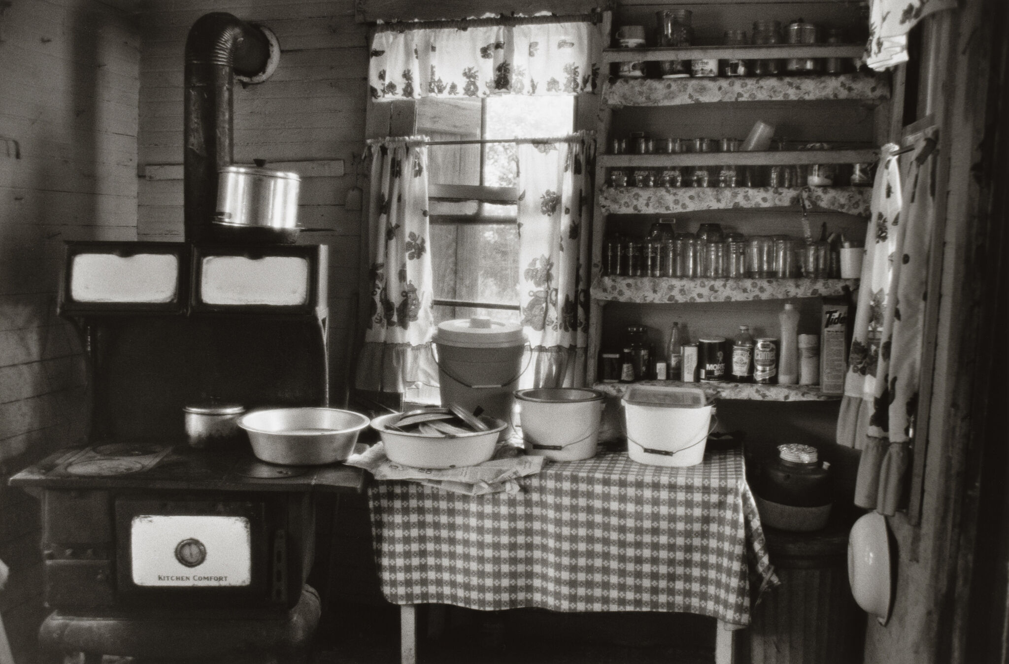 Vintage kitchen with a wood stove, pots, and a table covered in a checkered cloth. Shelves hold jars and cans, and floral curtains hang by the window.