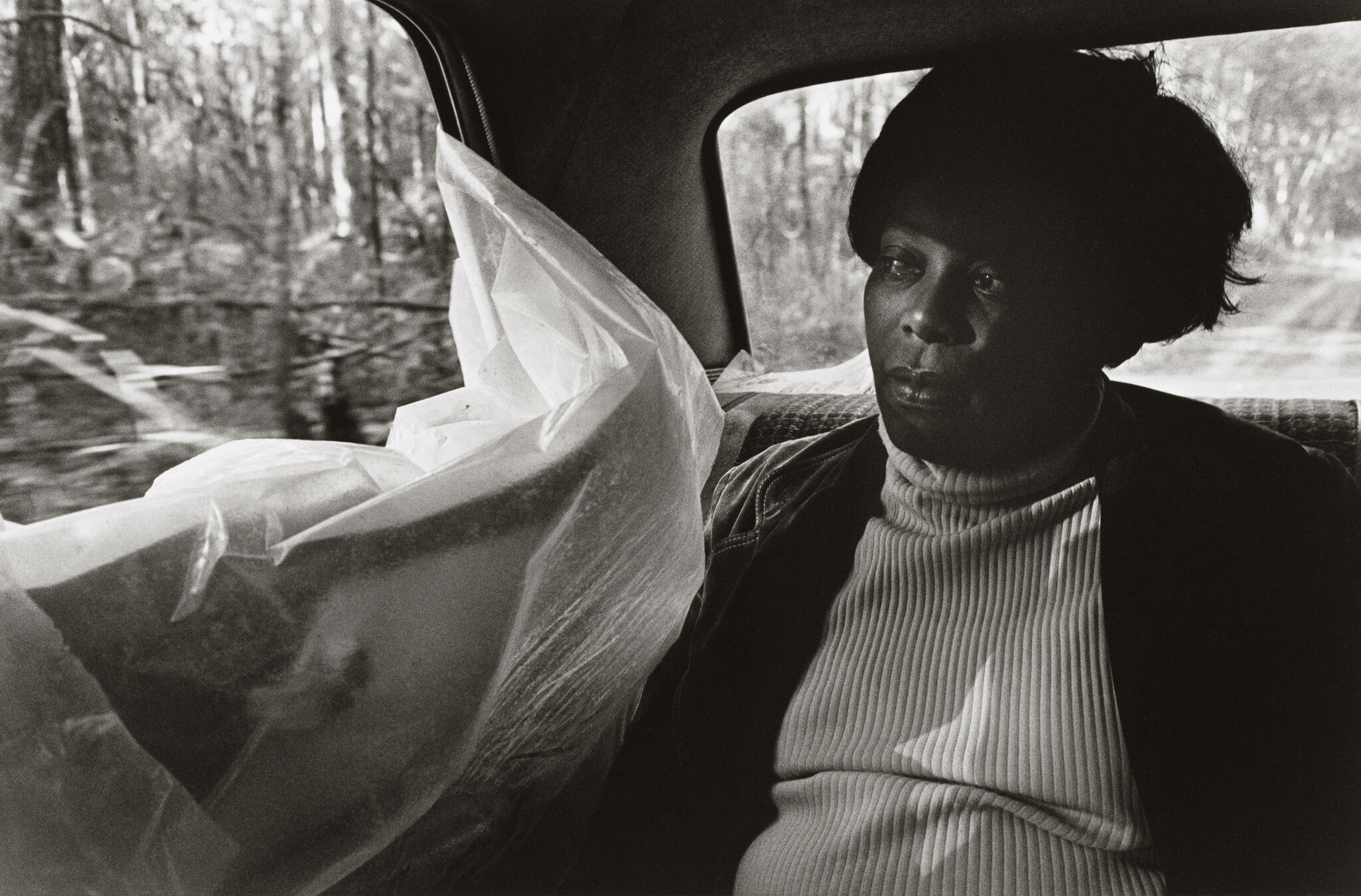 A woman in a turtleneck sits in the backseat of a car, gazing out the window. A plastic sheet partially covers the window.