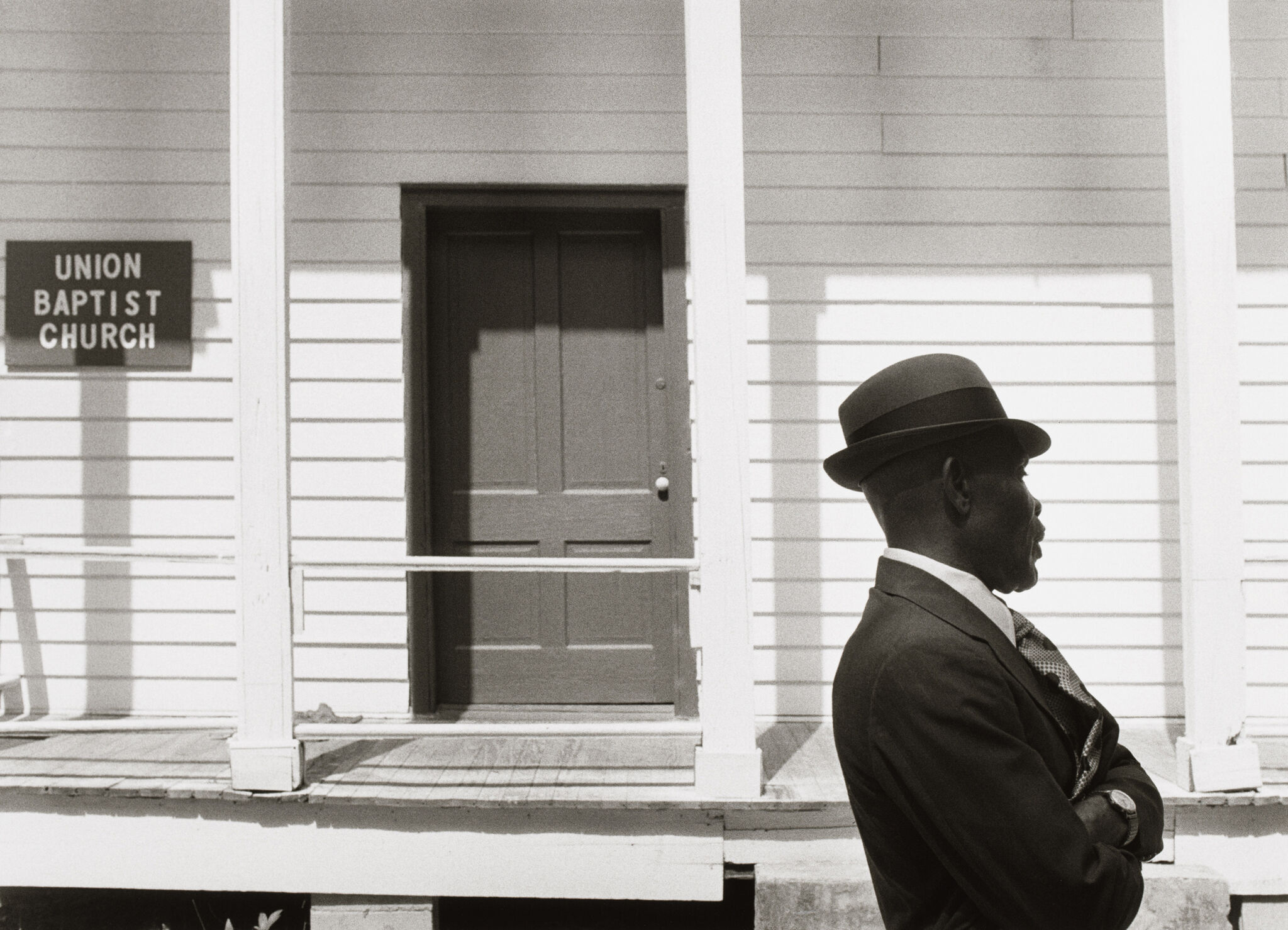 Man in a suit and hat stands in front of Union Baptist Church, a white wooden building with a closed door.