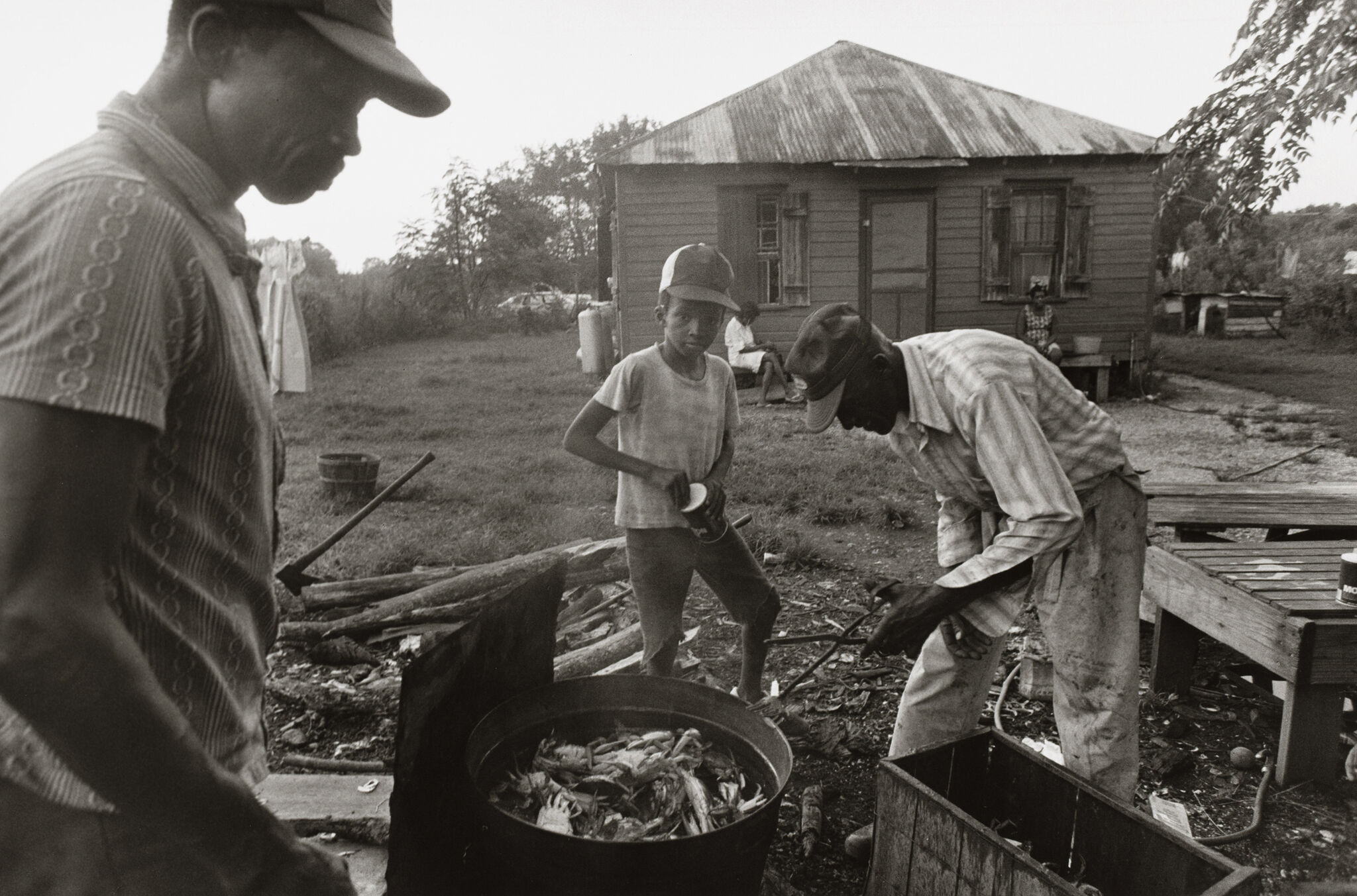 Three people cooking crabs outdoors near a rustic house, with one person sitting on the porch in the background.