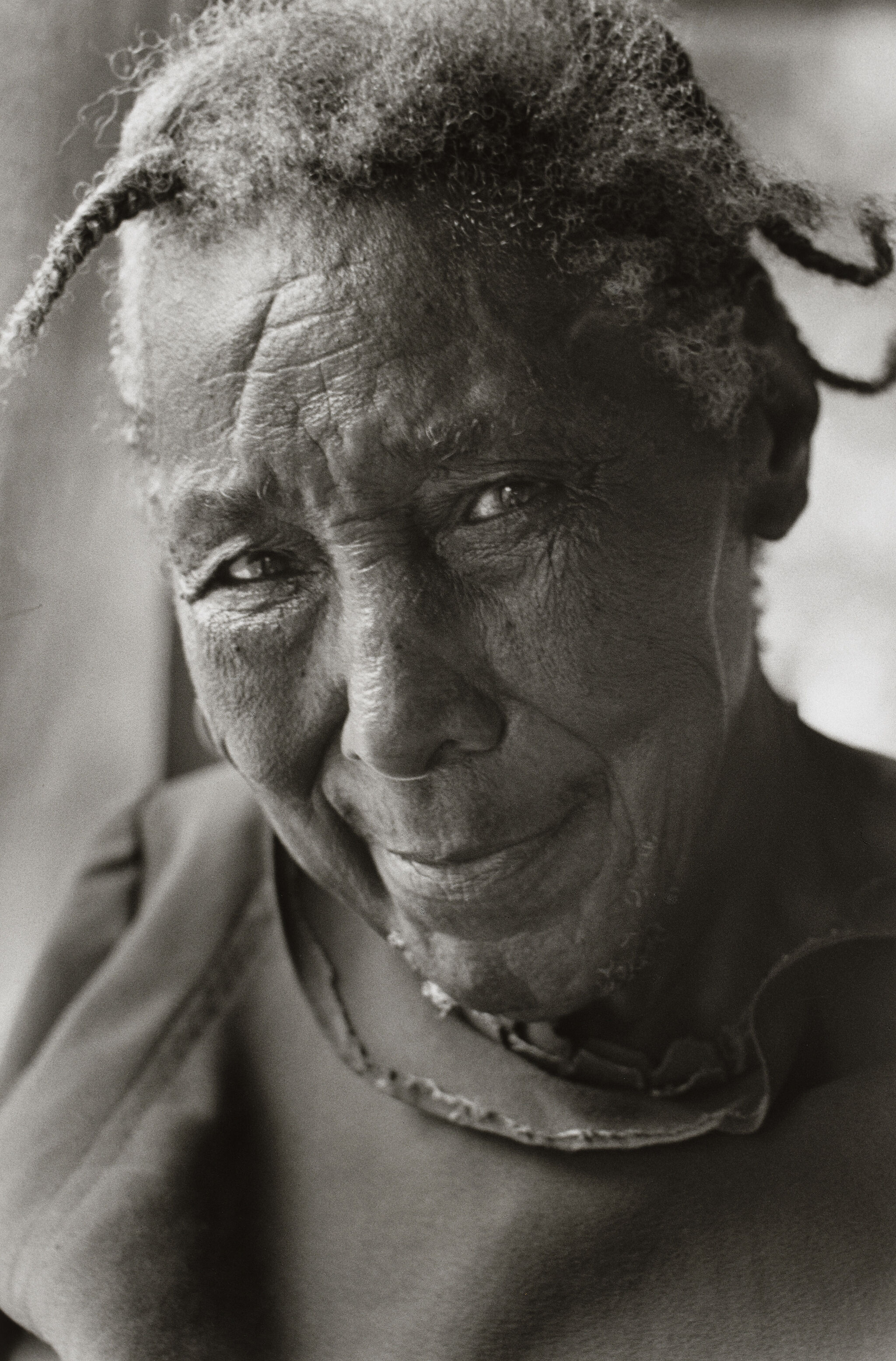 Elderly person with a gentle smile, wearing a simple garment. The black and white photo highlights their expressive eyes and textured skin.