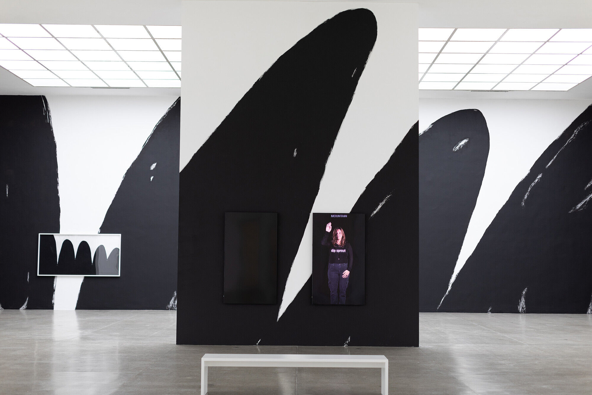 Gallery with abstract black and white wall art, two digital screens, and a white bench on a polished concrete floor.