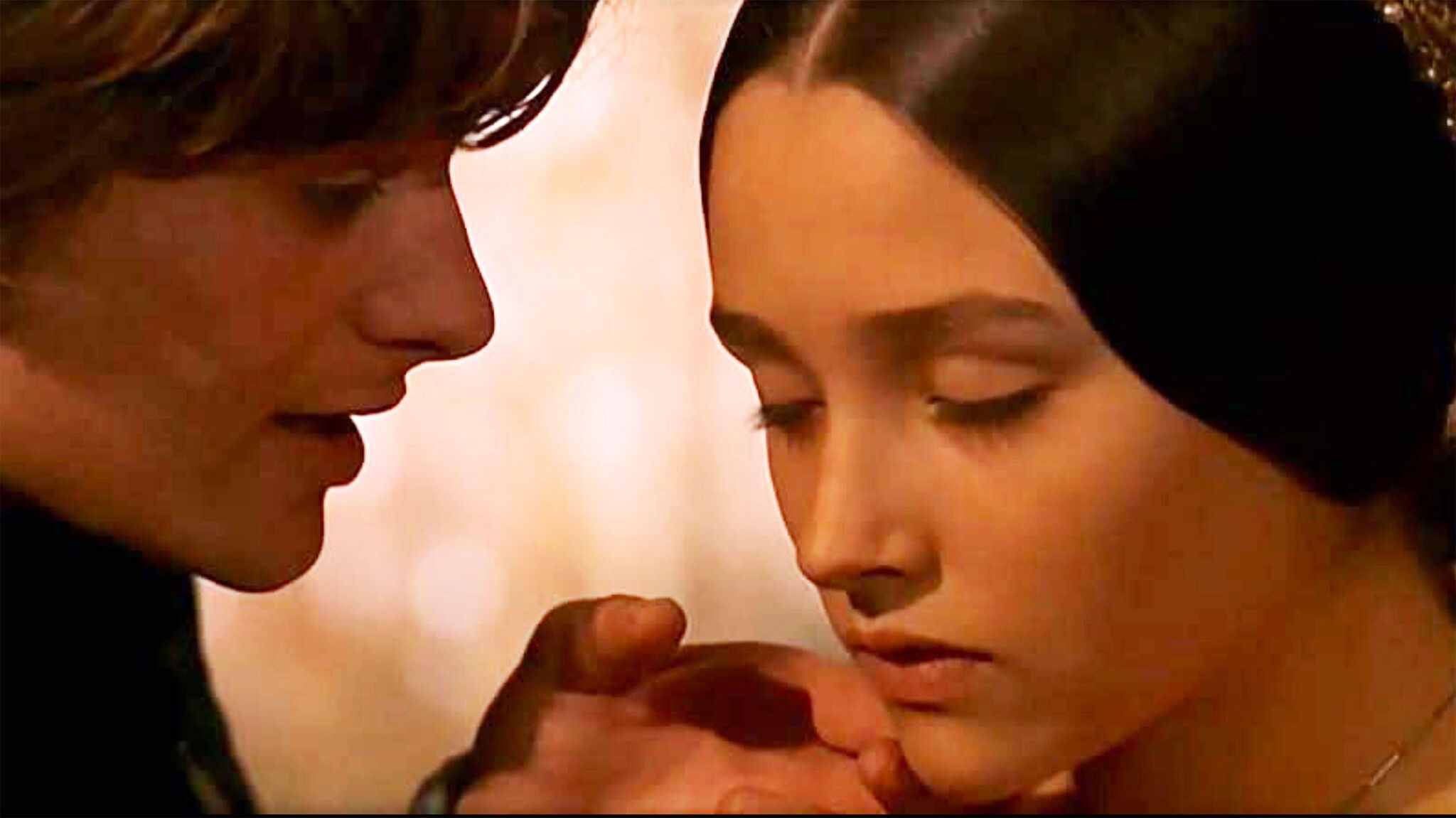 A young couple embraces on a stone balcony, surrounded by lush greenery, with a look of longing and concern.
