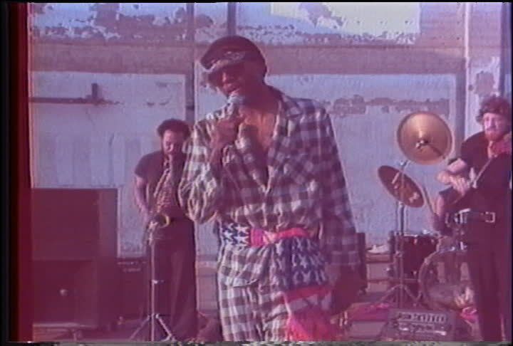 Man in a checkered suit and American flag scarf sings into a microphone. Band with saxophonist and drummer plays in the background.