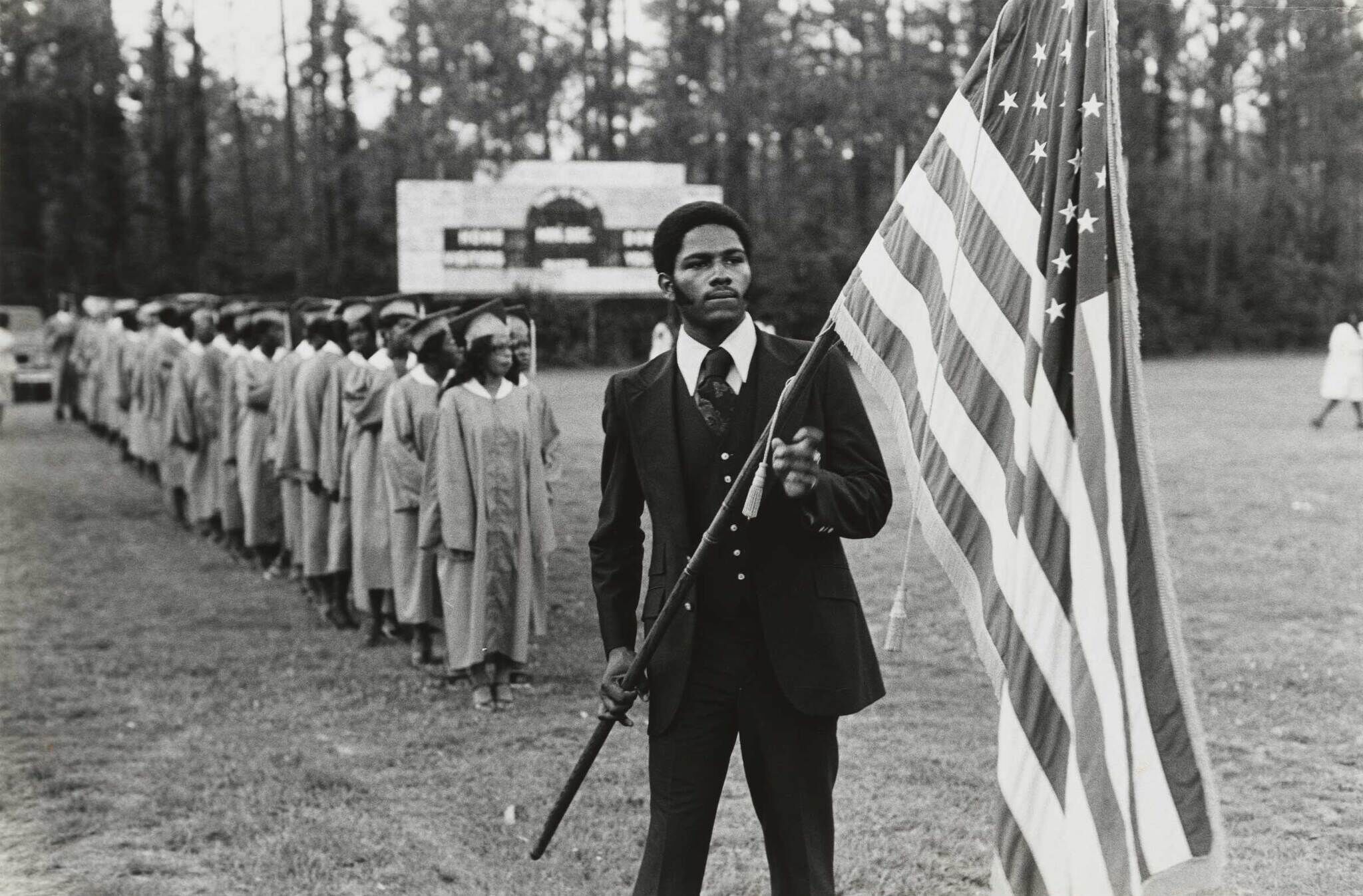 A man in a suit holds an American flag, leading a line of graduates in caps and gowns on a grassy field.