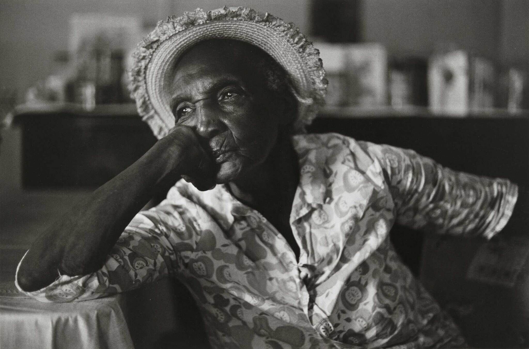 Elderly woman in a patterned dress and straw hat, resting her chin on her hand, gazing thoughtfully into the distance.