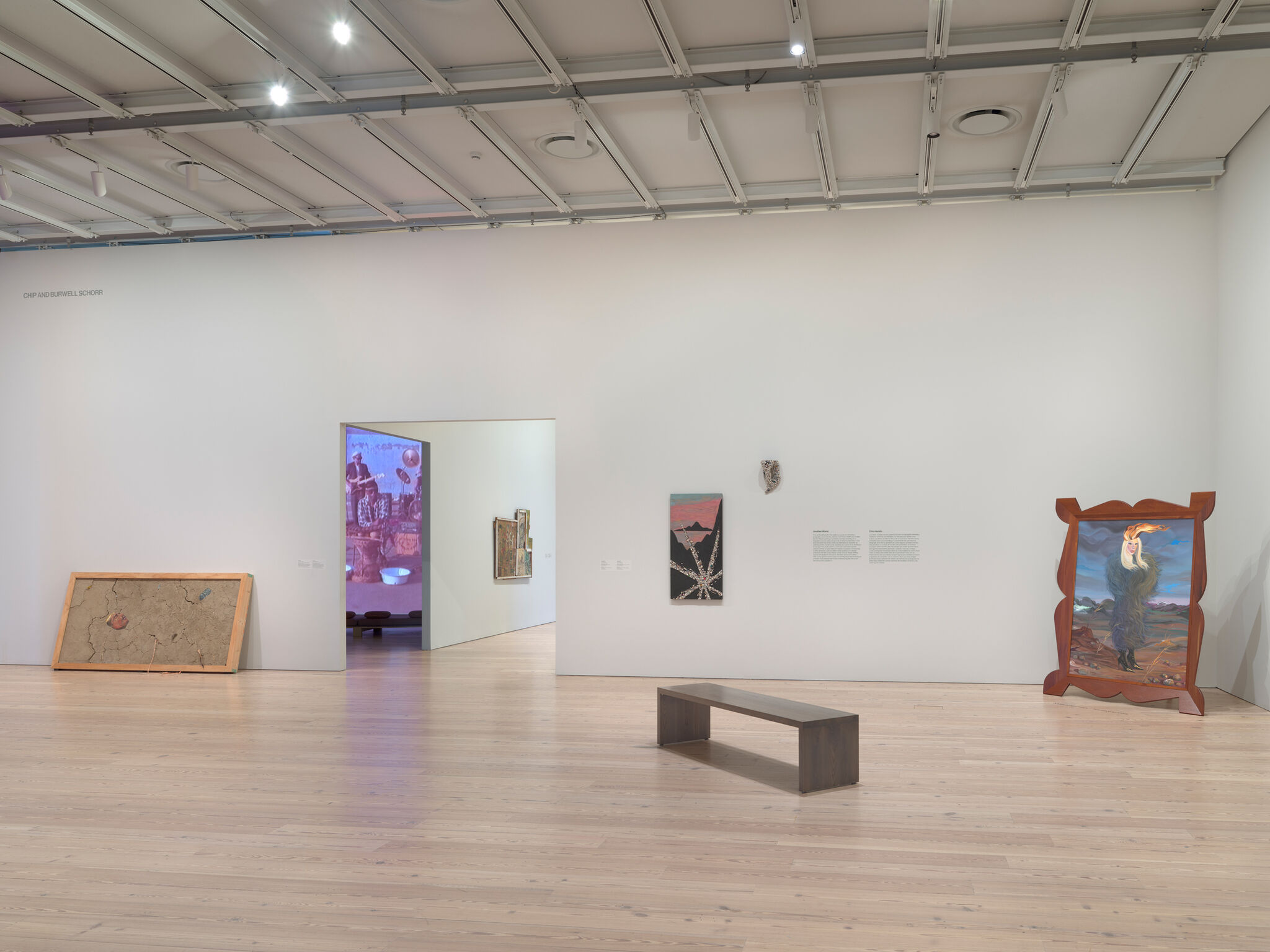 Art gallery with various paintings on white walls, a wooden bench in the center, and an open doorway leading to another room.