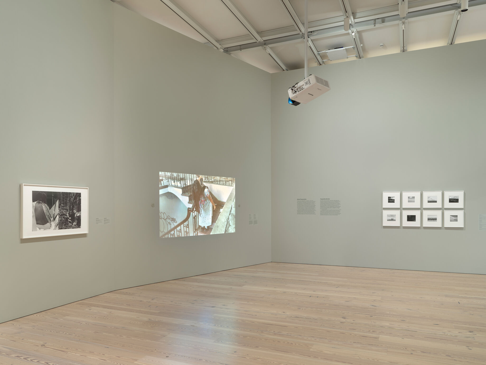 Gallery room with framed photos on walls and a video projection showing a person in a bathtub. Ceiling-mounted projector visible.