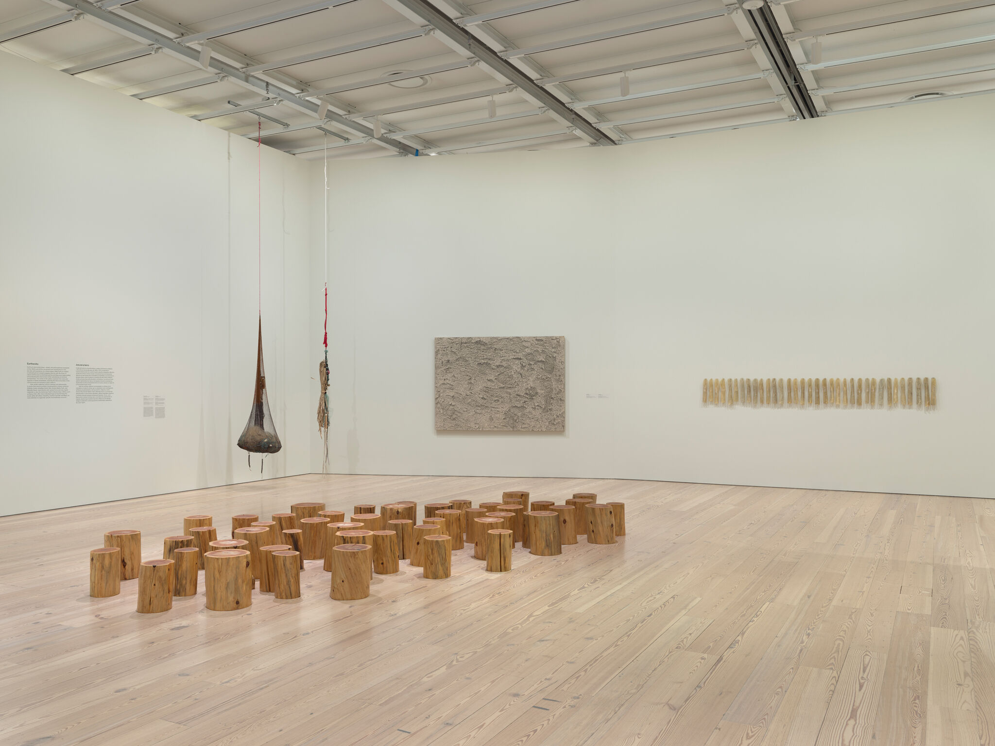 Art gallery with wooden stump sculptures on the floor, a hanging installation, and textured artworks on the walls.