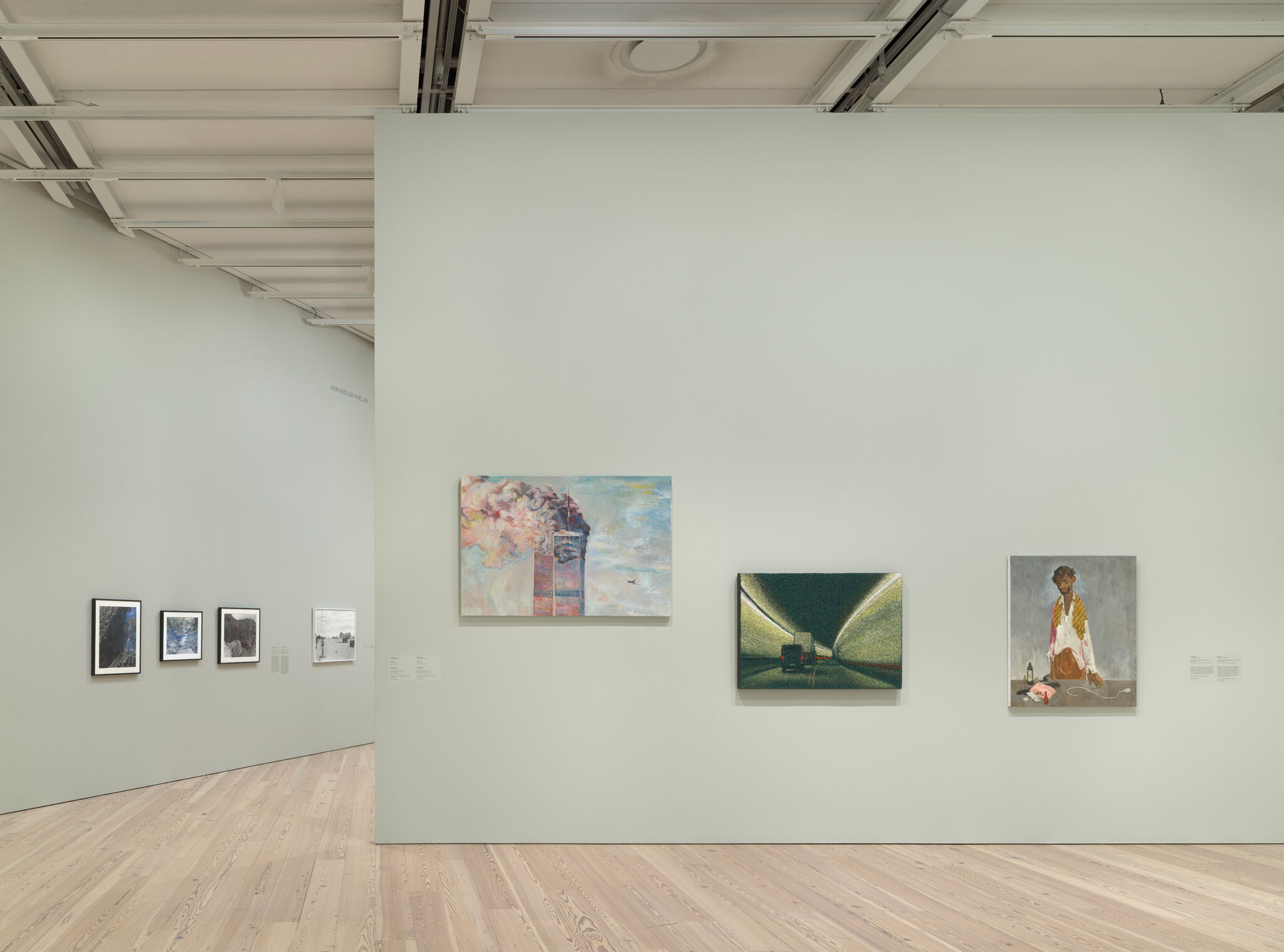 Art gallery with paintings on light green walls, featuring a smokestack, a tunnel, and a portrait. Light wood flooring and high ceiling.