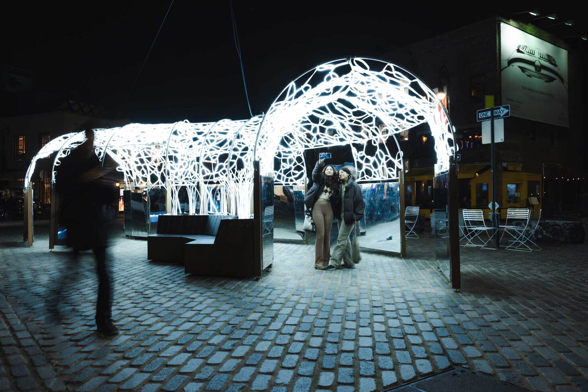 People take a selfie under a glowing, intricate light installation at night, with a blurred figure walking by on a cobblestone street.