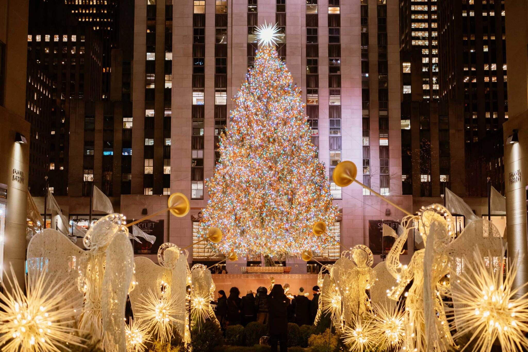 A large Christmas tree adorned with colorful lights stands in a city plaza, surrounded by illuminated angel decorations and a crowd of onlookers.