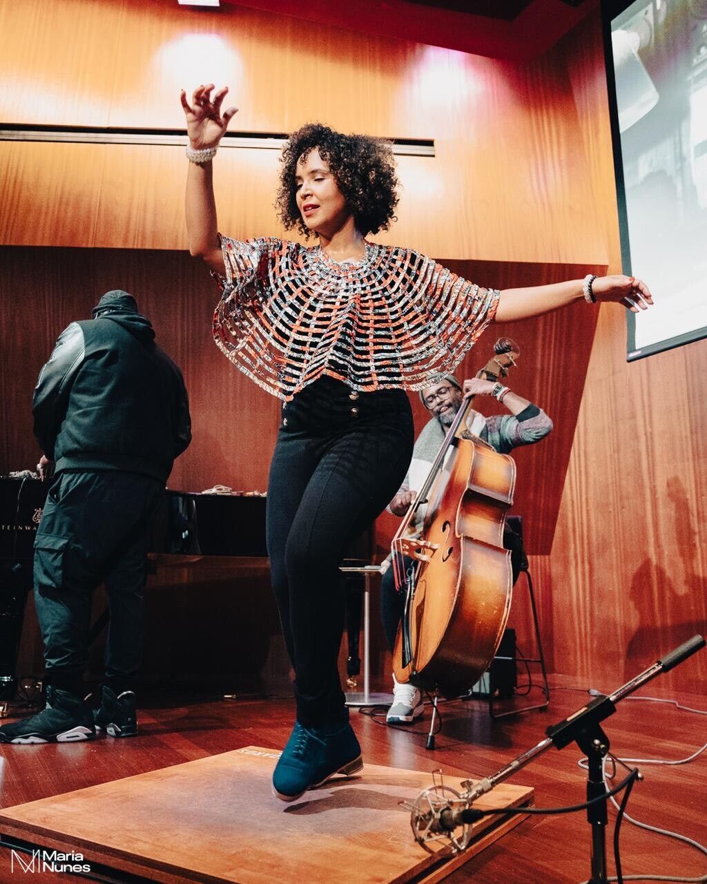 Alt text: Lisa La Touche, a tap dancer, performs energetically on a wooden platform wearing a colorful, web-patterned top and dark pants. Her arms are outstretched gracefully as she moves with precision and rhythm. Behind her, a musician plays an upright bass, and another individual is at the piano, set against a warm wooden backdrop. A microphone is positioned near her feet to capture the sound of her tapping.