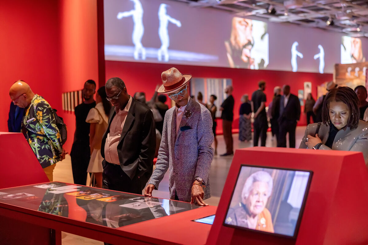 In forefront, four visitors explore a glass case and mounted screens displaying archival footage. In background, many people explore galleries surrounded by large video installation.