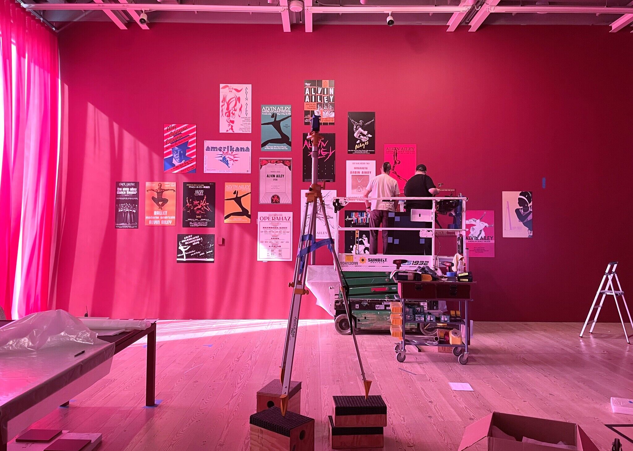 Two people on a lift arrange colorful Alvin Ailey posters on a red wall in a gallery, with tools and equipment scattered on the wooden floor.