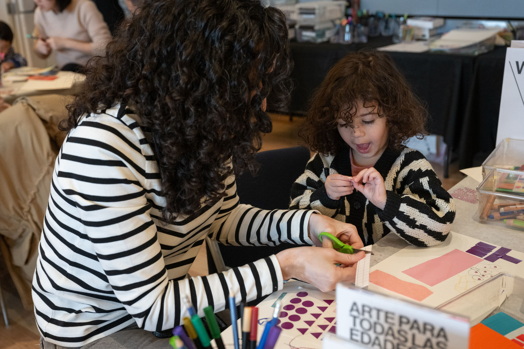 A family is creating art together during Free Second Sunday at the Whitney Museum. An adult with curly hair and a striped sweater is helping a young child with curly hair work on an art project. They are seated at a table filled with colorful supplies, including markers, paper, and scissors. A sign in the foreground reads, "Arte para todas las edades," highlighting that the activity is designed for all ages.