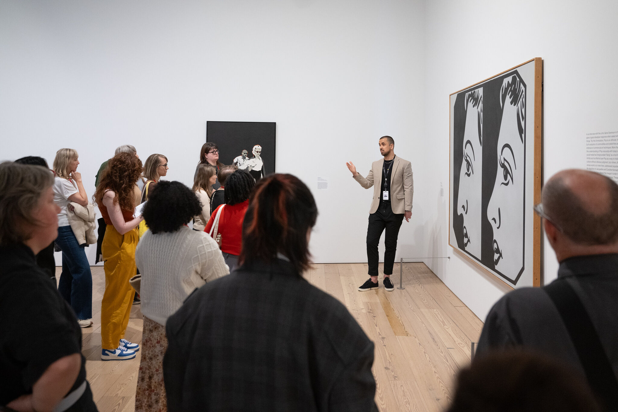 A man in a tan blazer raises his hand bent at the elbow toward group of onlookers in front of a painting of a simplified, cartoon-like, double portrait in black and white.