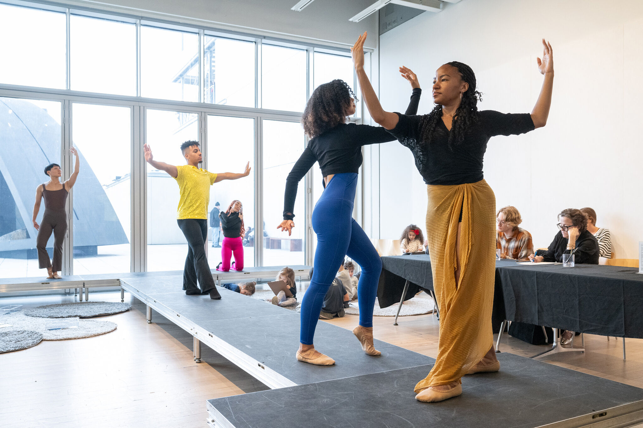 Dancers perform on a stage in front of a panel of judges, with large windows in the background.