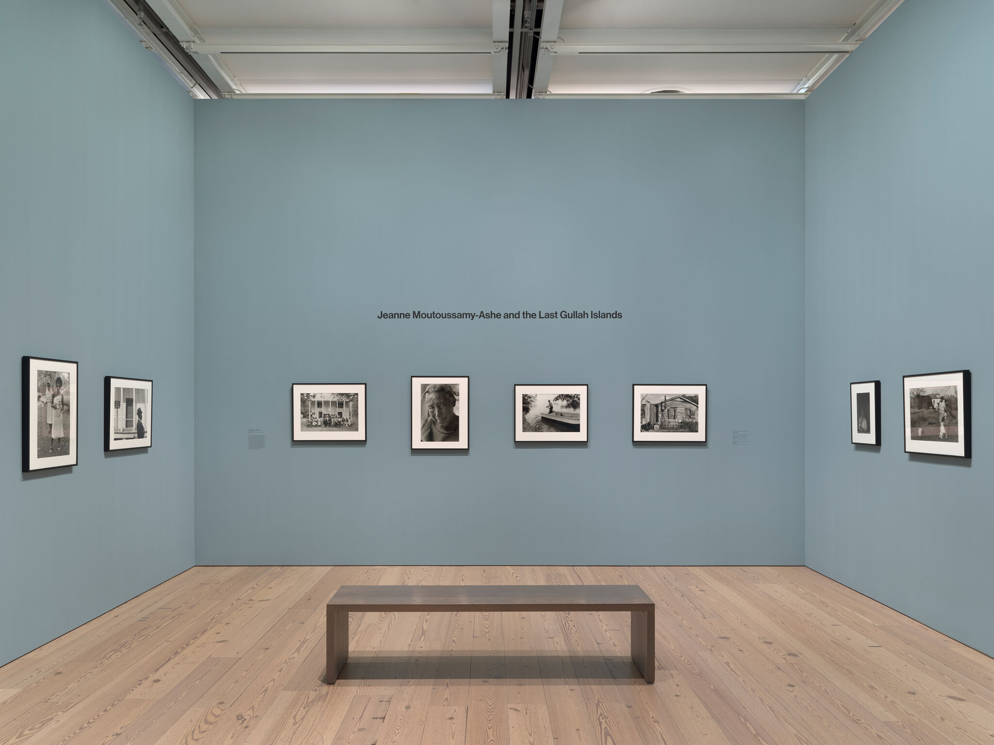 Gallery room with blue walls displaying black-and-white photos by Jeanne Moutoussamy-Ashe, titled "The Last Gullah Islands," with a bench.