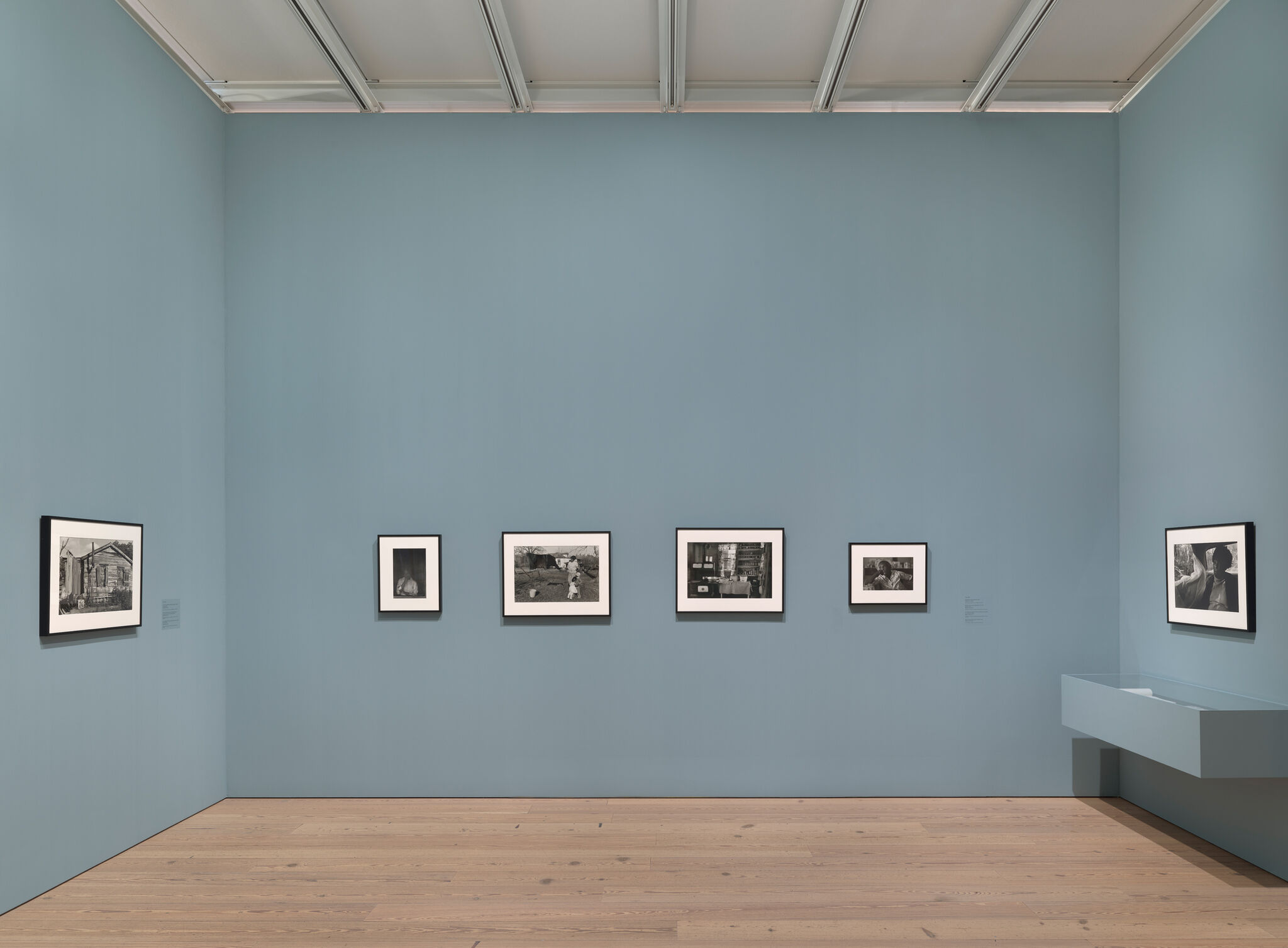 Gallery with six framed black-and-white photos on a light blue wall, wooden floor, and a small shelf on the right.