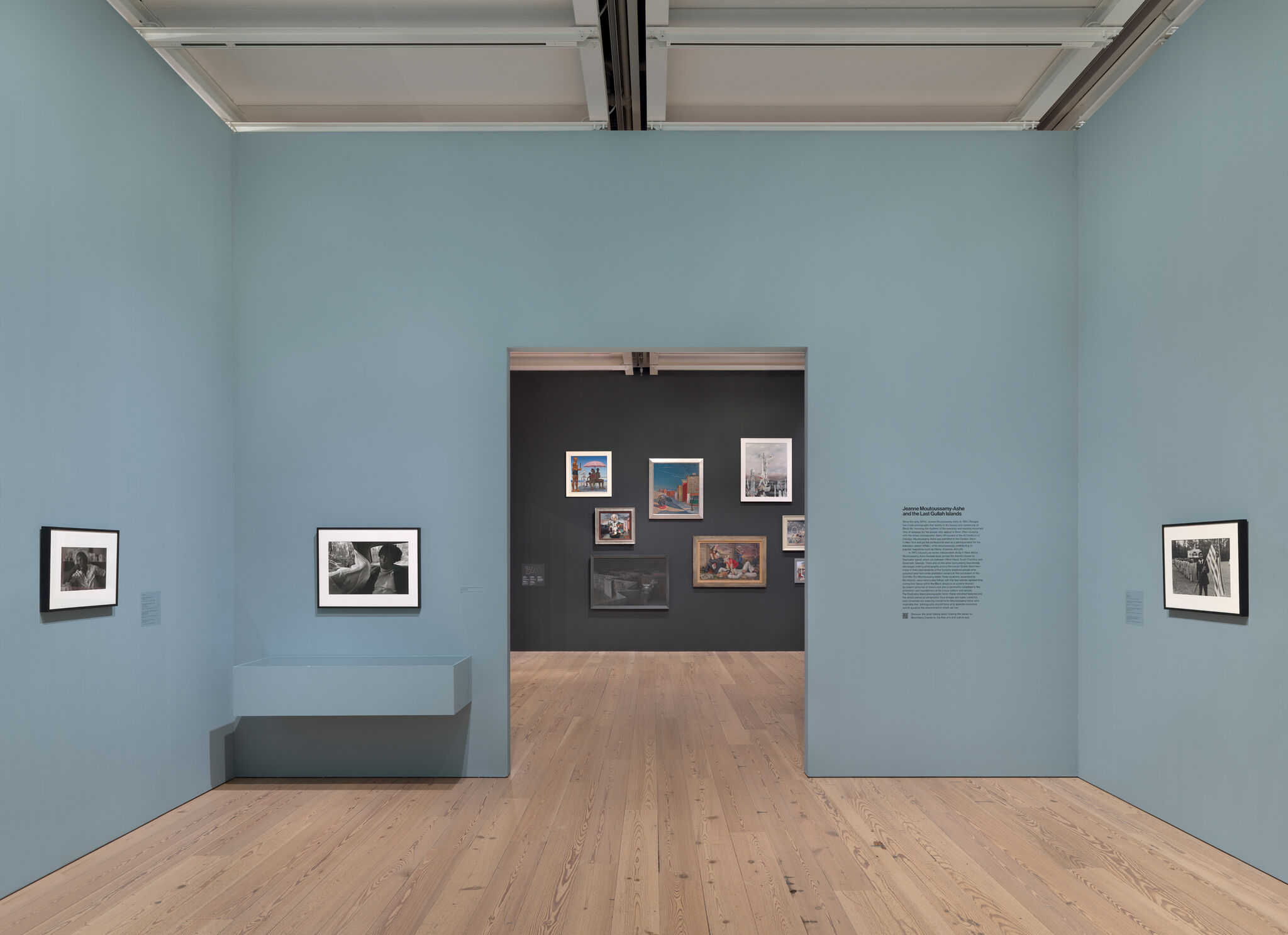 Art gallery with light blue walls, featuring black-and-white photos. A doorway leads to a darker room with colorful framed artworks.