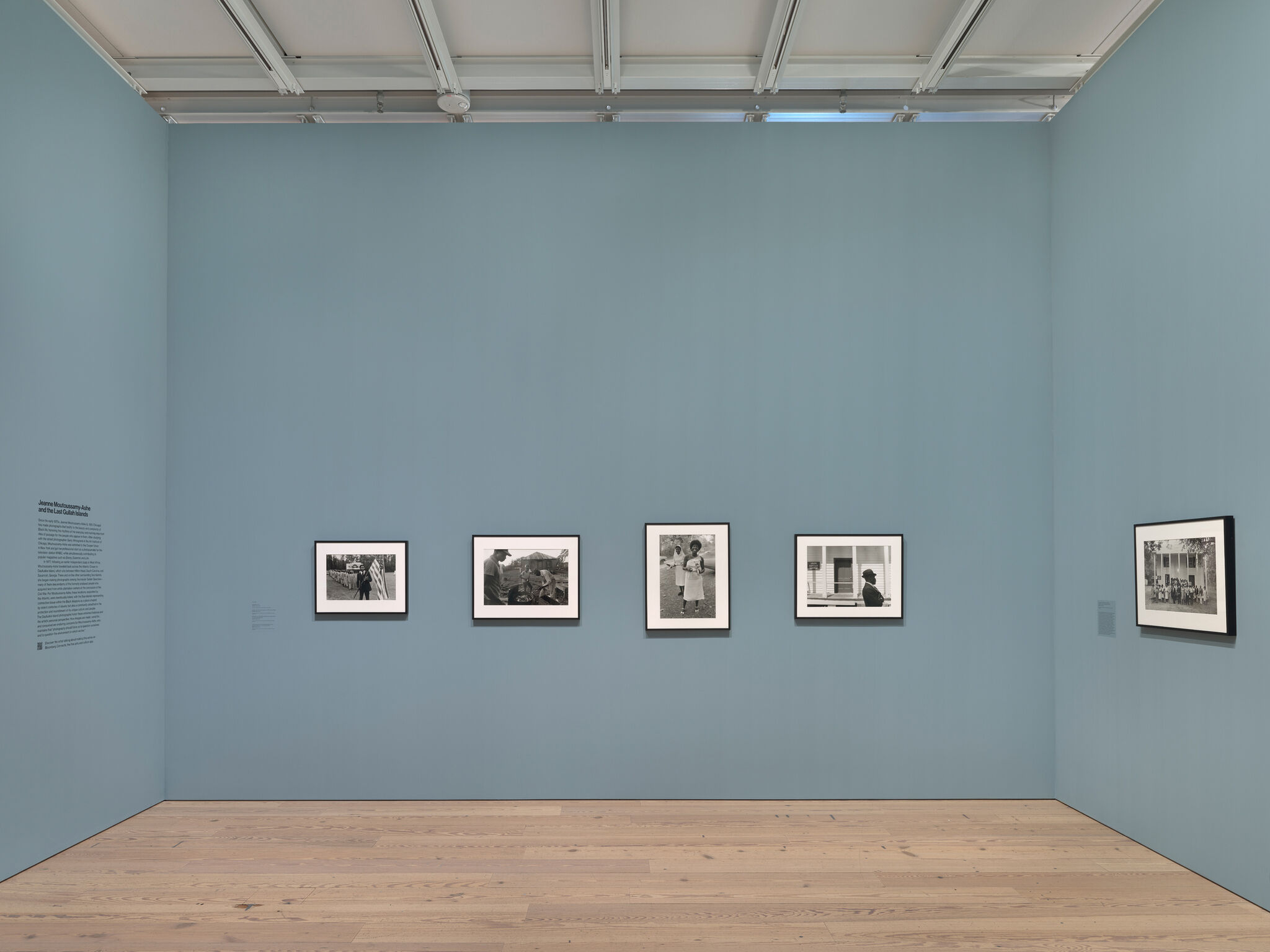 Gallery with five framed black-and-white photographs on a light blue wall, wooden floor, and overhead lighting.